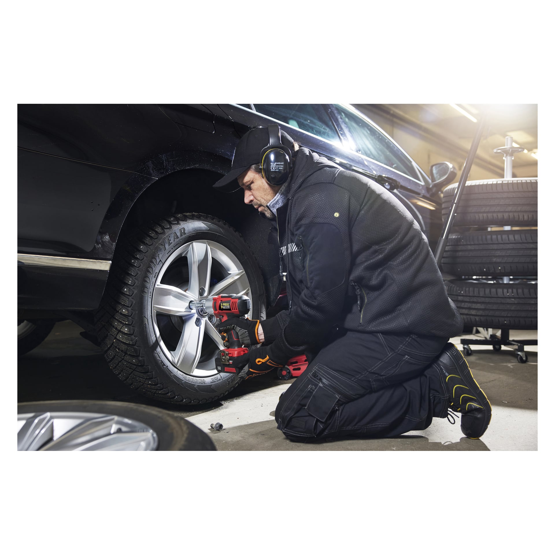 Kuulokkeet ja työkäsineet päässä oleva henkilö polvistuu mustan auton viereen autotallissa, käyttäen Meec Tools Multiseries -paristokäyttöistä törmäysavainta IW-18V EASY 100 renkaan vaihtoon. Taustalla on useita renkaita.