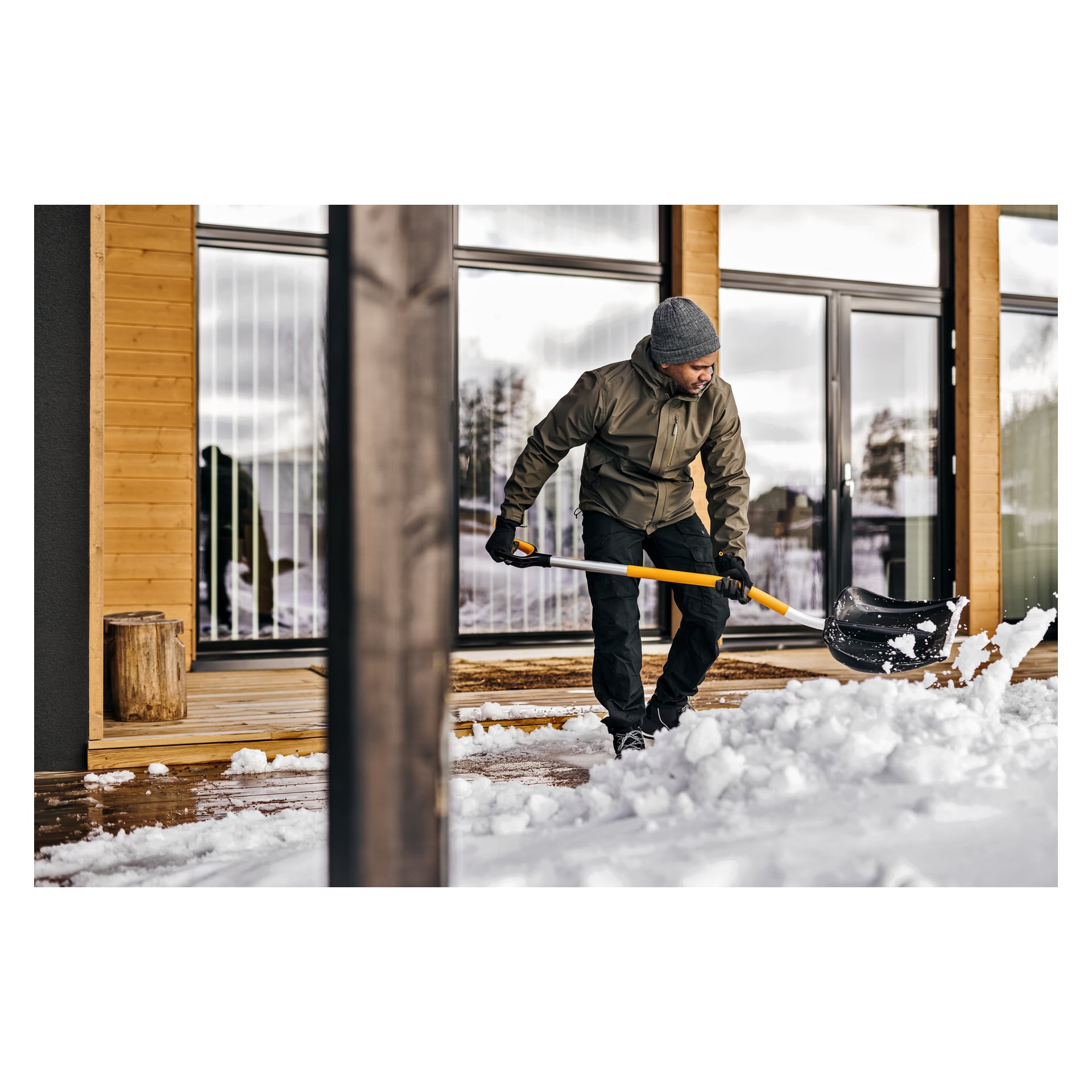 Osoba ubrana w zimowe ubrania, w tym kurtkę, kapelusz i rękawiczki, odśnieża śnieg z drewnianego tarasu za pomocą Fiskars Ergonomic X-Series przed nowoczesnym budynkiem z dużymi oknami.