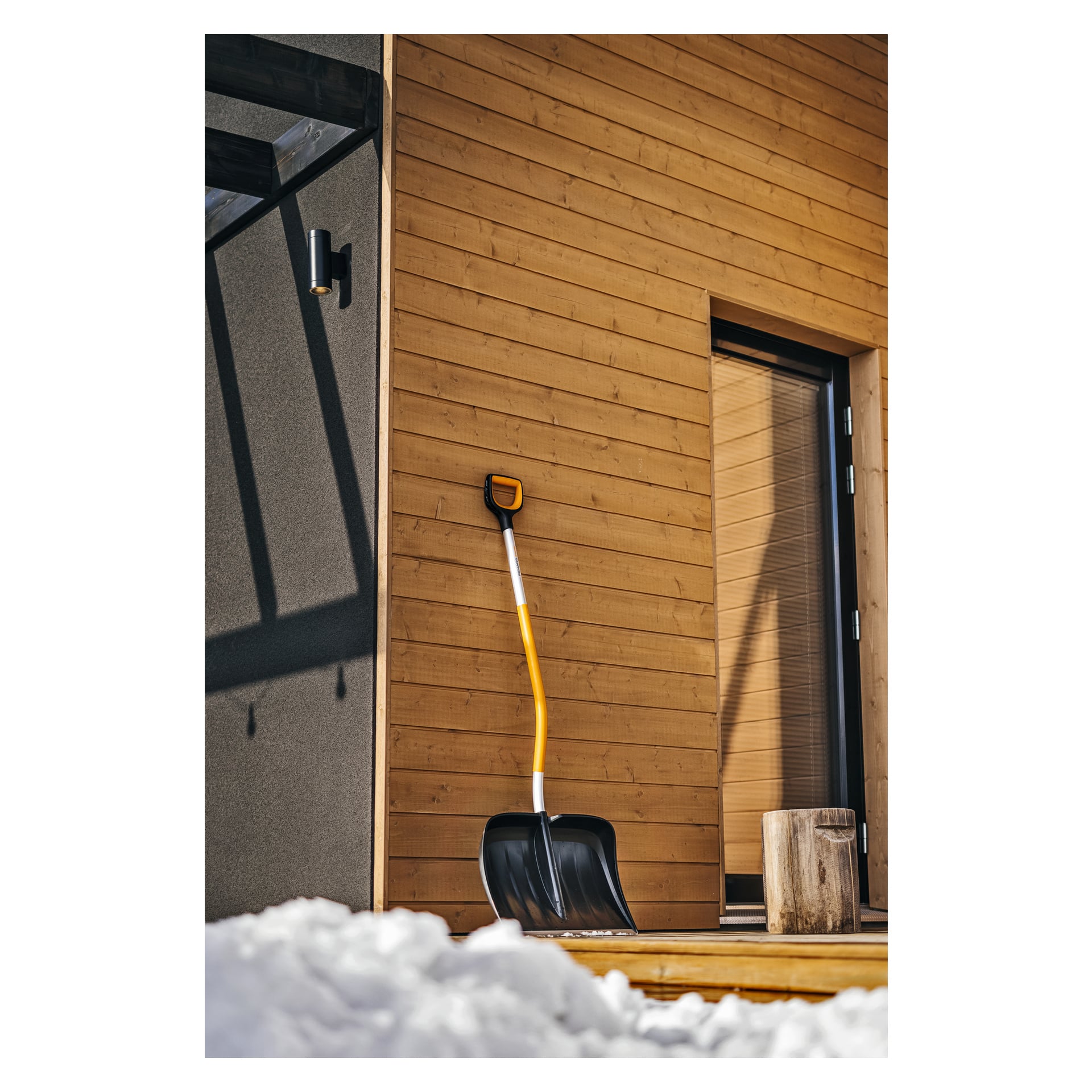 Żółtorękowa łopata Fiskars Ergonomic X-Series opiera się o drewnianą ścianę domu przy drzwiach, z małym pniem drzewa w pobliżu i śniegiem pokrywającym ziemię przed nimi.