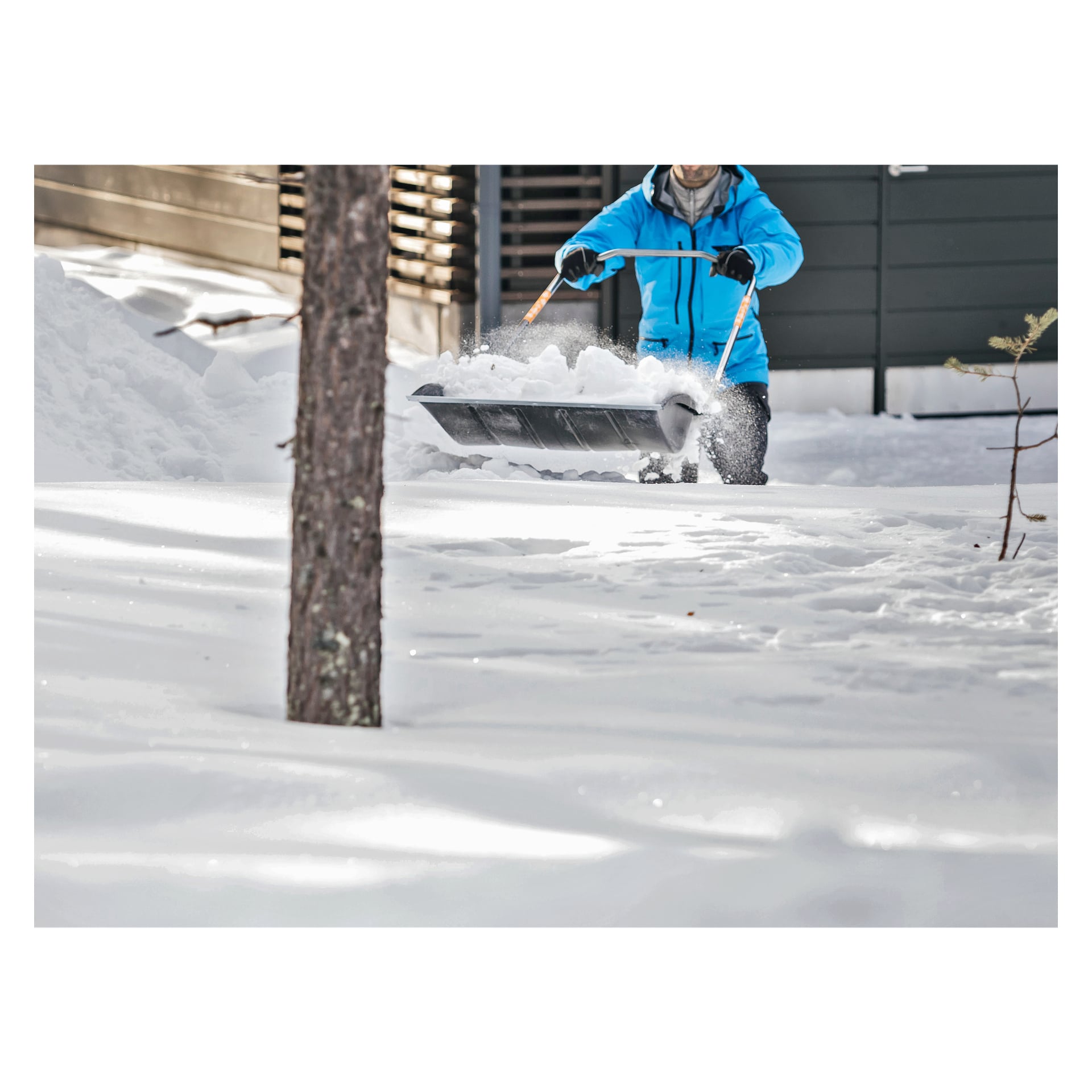 Iklädd en blå jacka skottar någon djup snö från en trädgård med Fiskars Sledge SnowXpert snöskyffel, som har en non-stick hink. Träd är synliga runt dem, och deras ansikte är vänt bort medan de rensar snön.