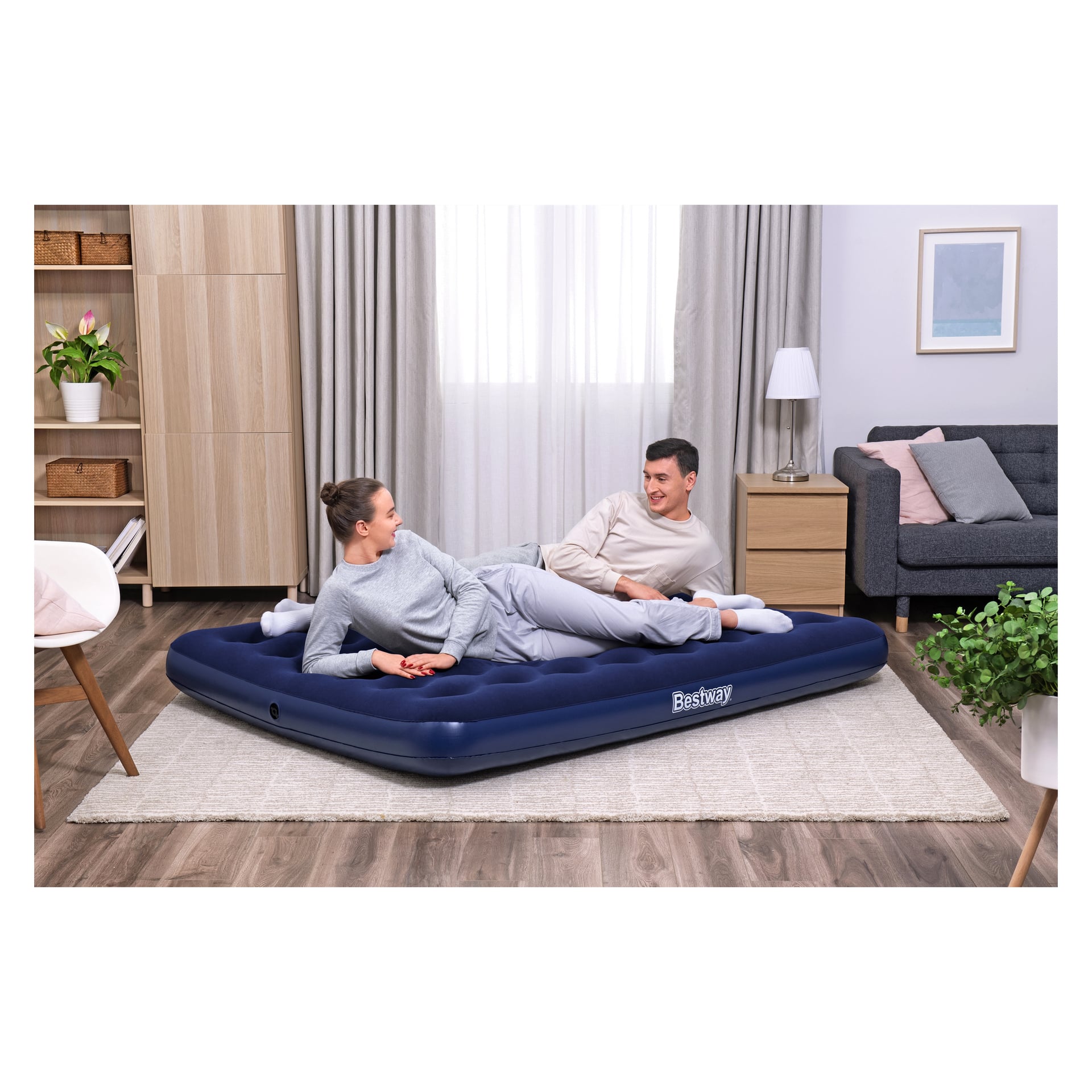 Två personer i myskläder ligger på en Bestway Inflatable Mattress Queen i ett vardagsrum med trägolv, möbler i neutrala färger, krukväxter och ett fönster med tunna gardiner. De står vända mot varandra och ser bekväma och avslappnade ut.