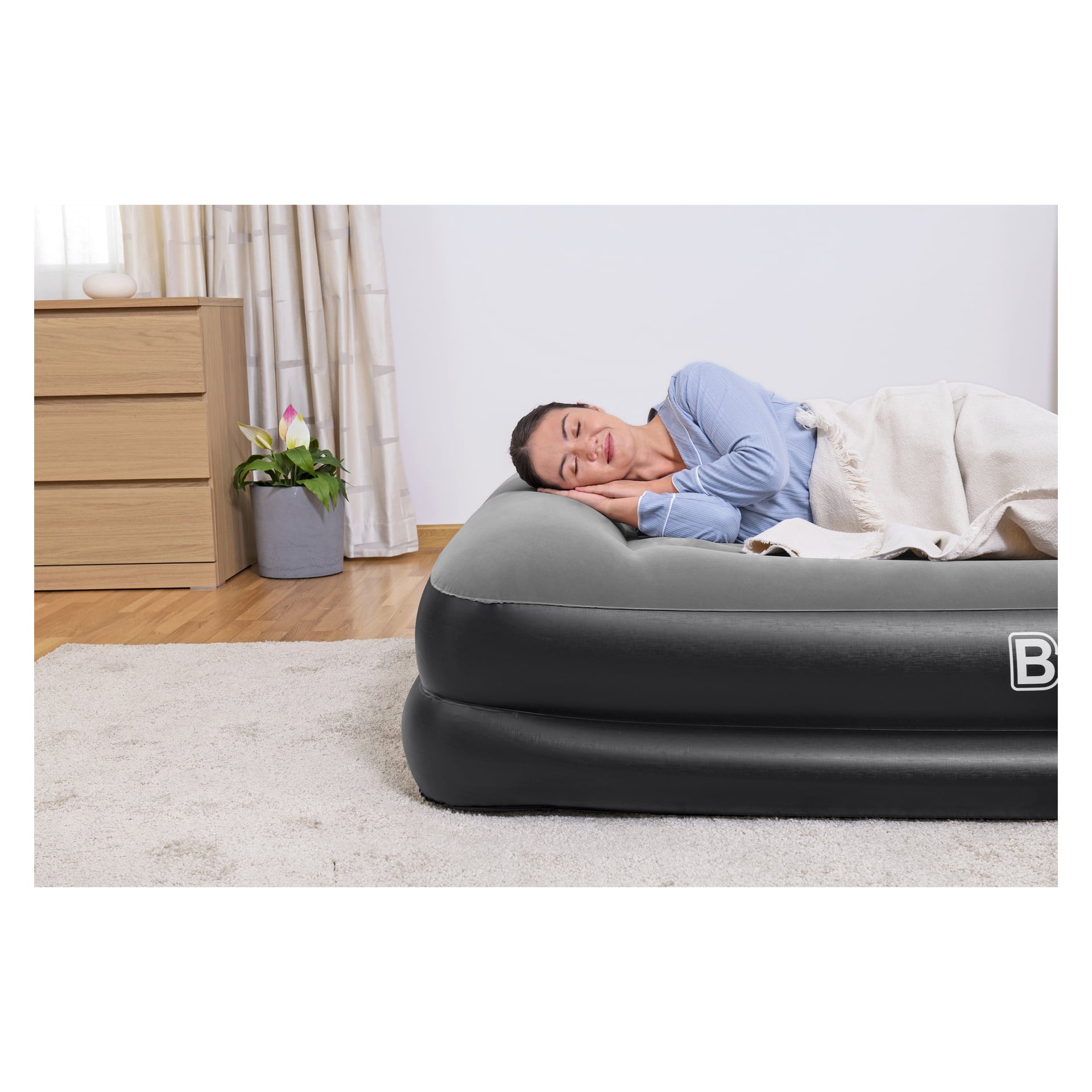 En kvinna i blå skjorta sover på Bestway Inflatable-madrassen med inbyggd pump Queen, täckt av en beige filt i ett prydligt sovrum med trämöbler, en växt och en ljus matta.