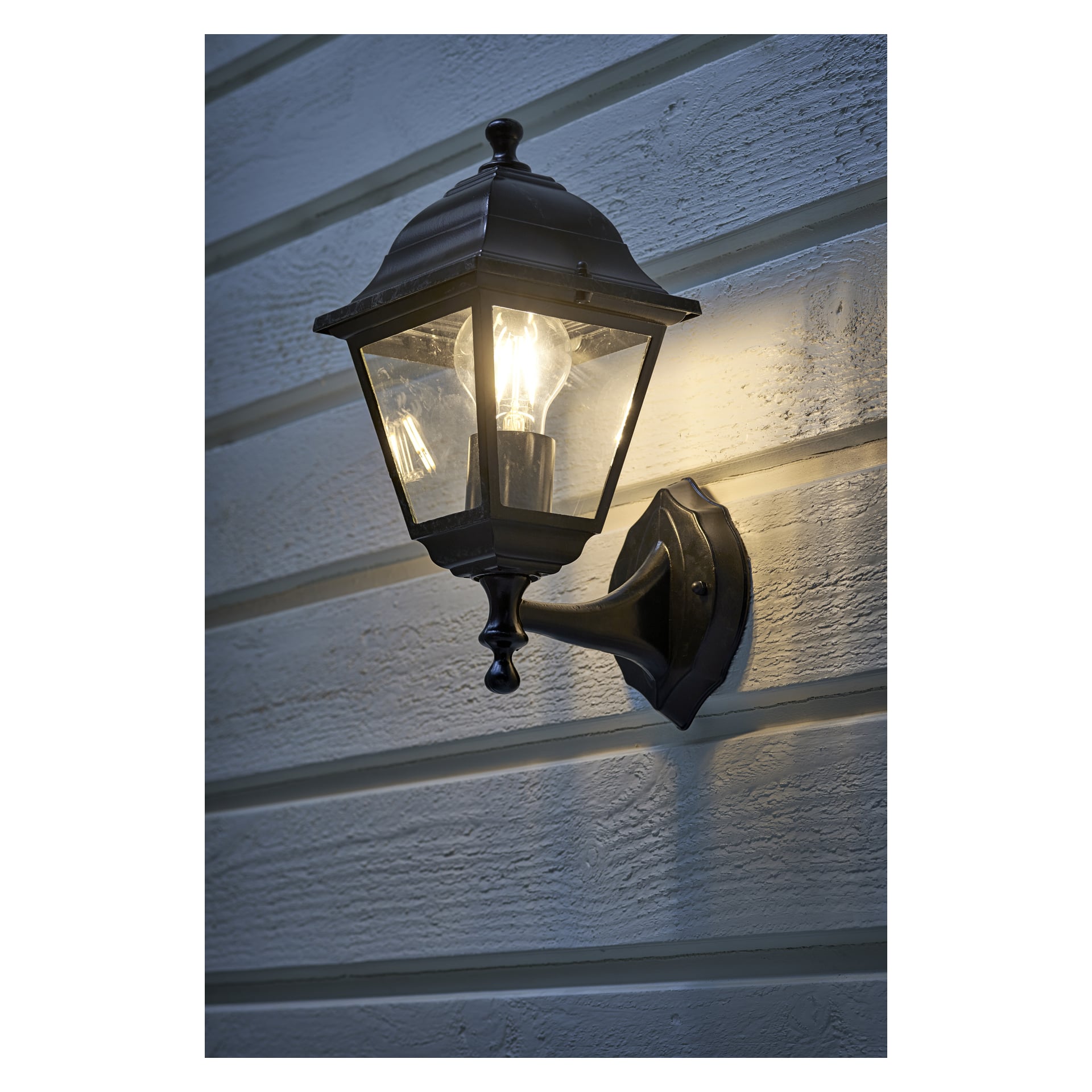 En svart metallvägglampa av EKVIP är fäst vid en ljus trävägg, där dess tända lampa skapar ett varmt sken och mjuka skuggor längs plankorna.