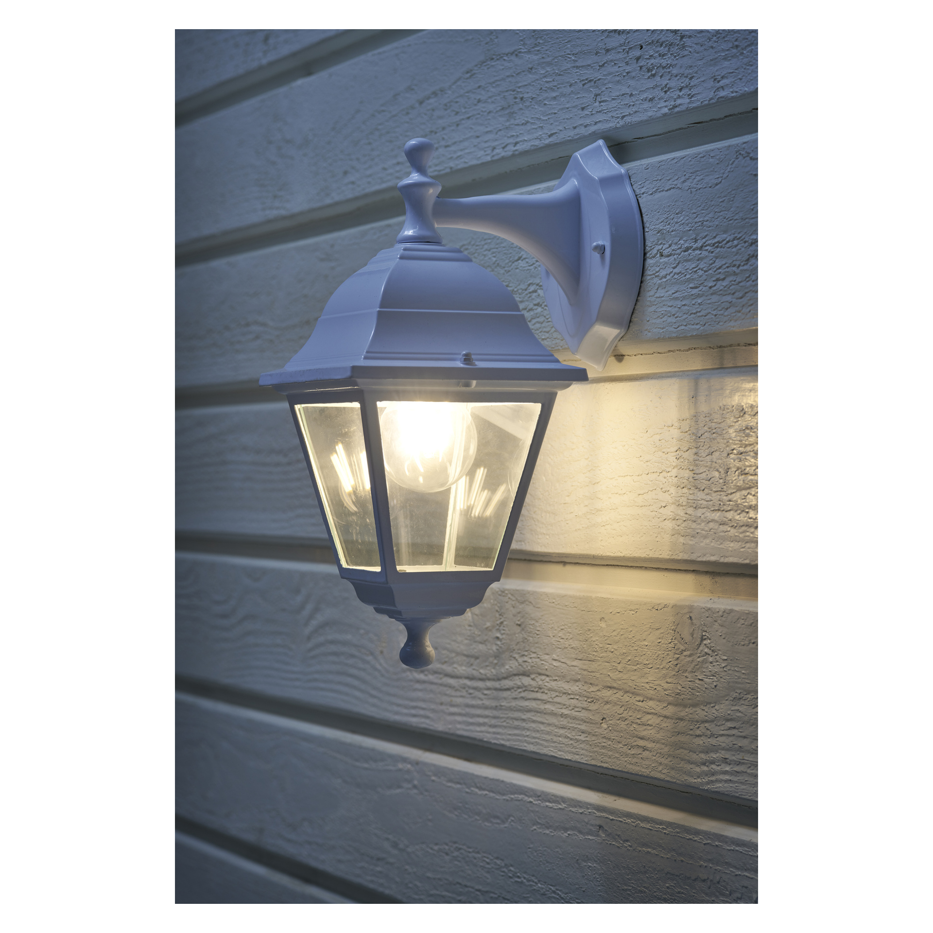 En klassisk EKVAP-vägglampa är fäst på ljus horisontell fasad, dess lampa exponerad och avger ett milt sken på väggen.