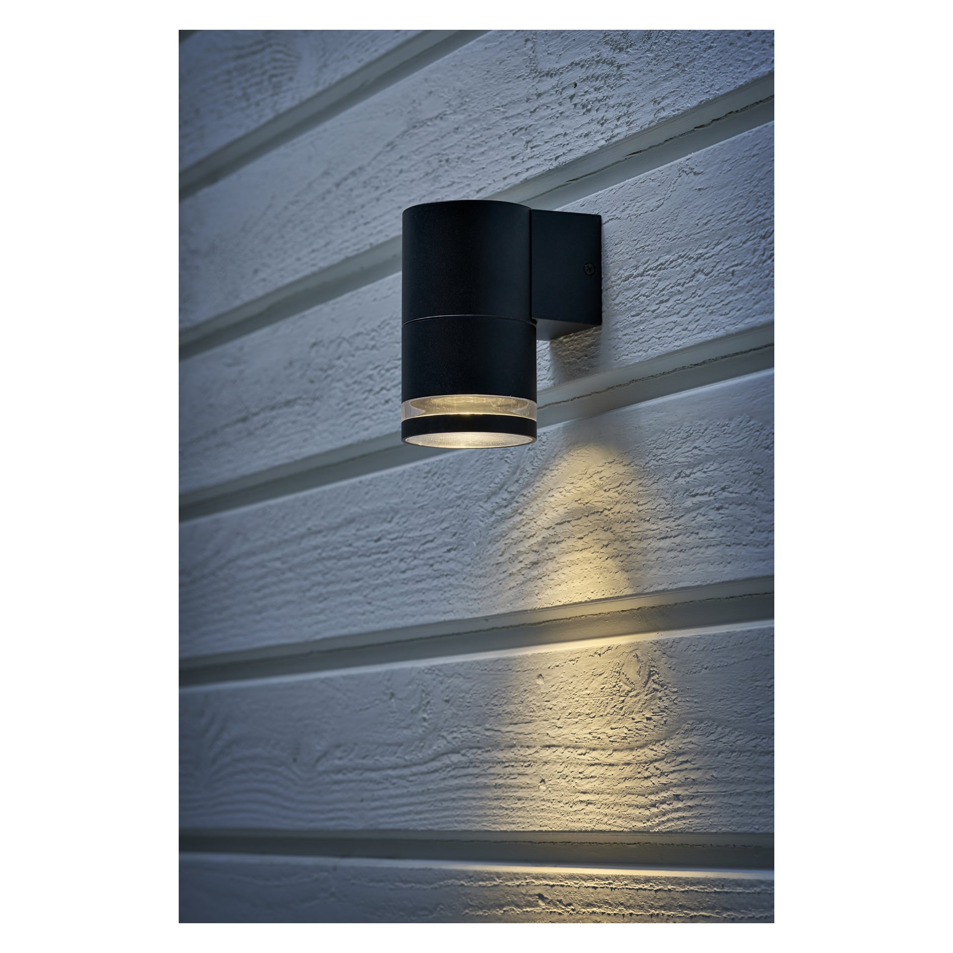 En svart EKVAP-väggstrålkastare med en slank cylindrisk form är fäst vid en vit träplankvägg. Den är påslagen och kastar ett milt nedåtriktat ljus som framhäver den texturerade väggytan under.