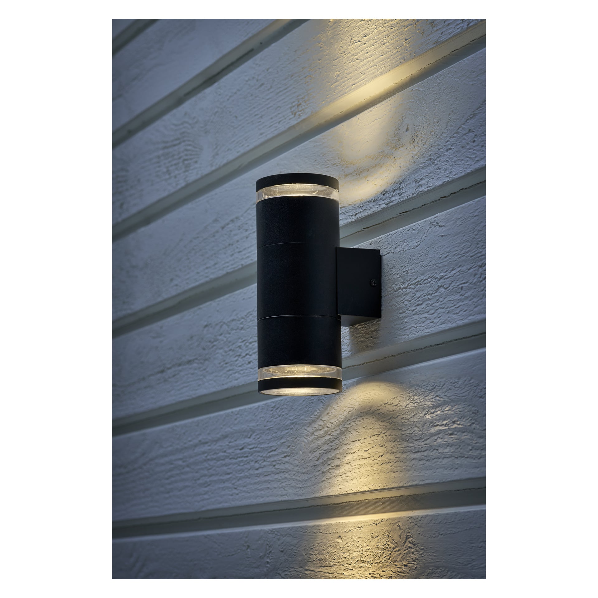 En svart cylindrisk EKVAP väggstrålkastare är installerad på en horisontellt panelad vägg, vilket kastar ljus både uppåt och nedåt och skapar cirkulära ljuspölar ovanför och under armaturen.