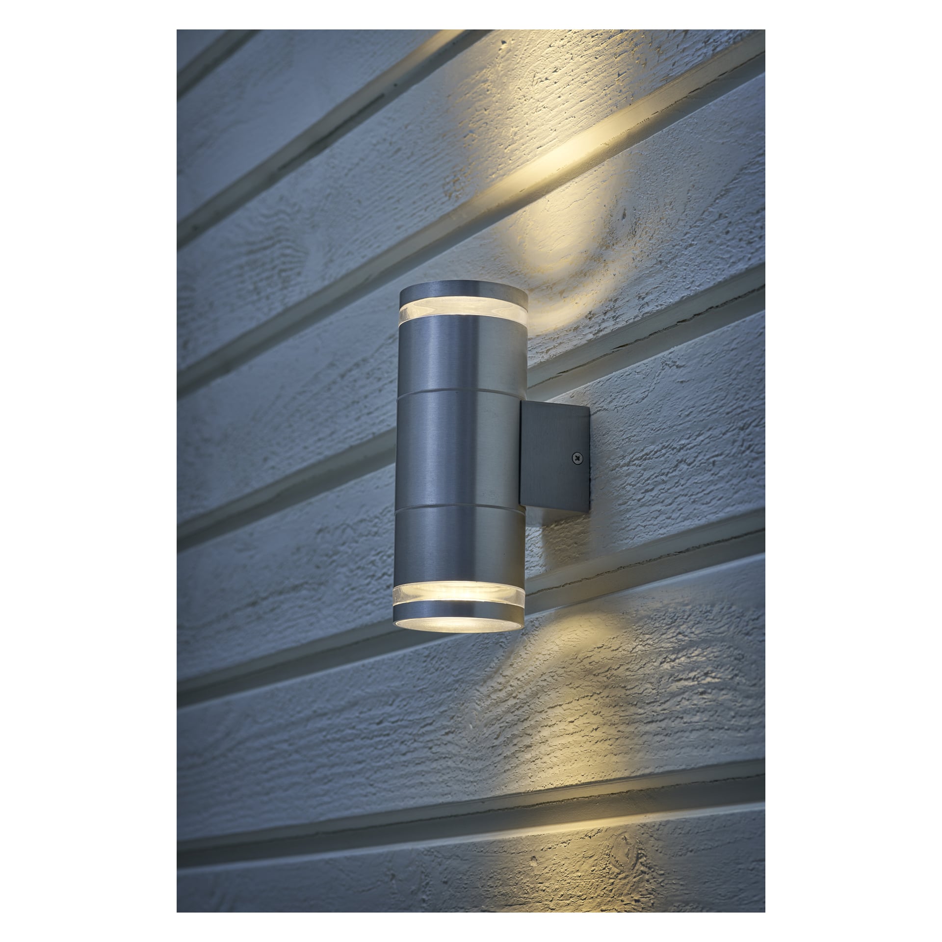 EKVIP Wall spotlight har en cylindrisk form av borstat metall och är monterad på horisontell fasad, vilket riktar ljuset både uppåt och nedåt för att skapa mjuka mönster på ytan.