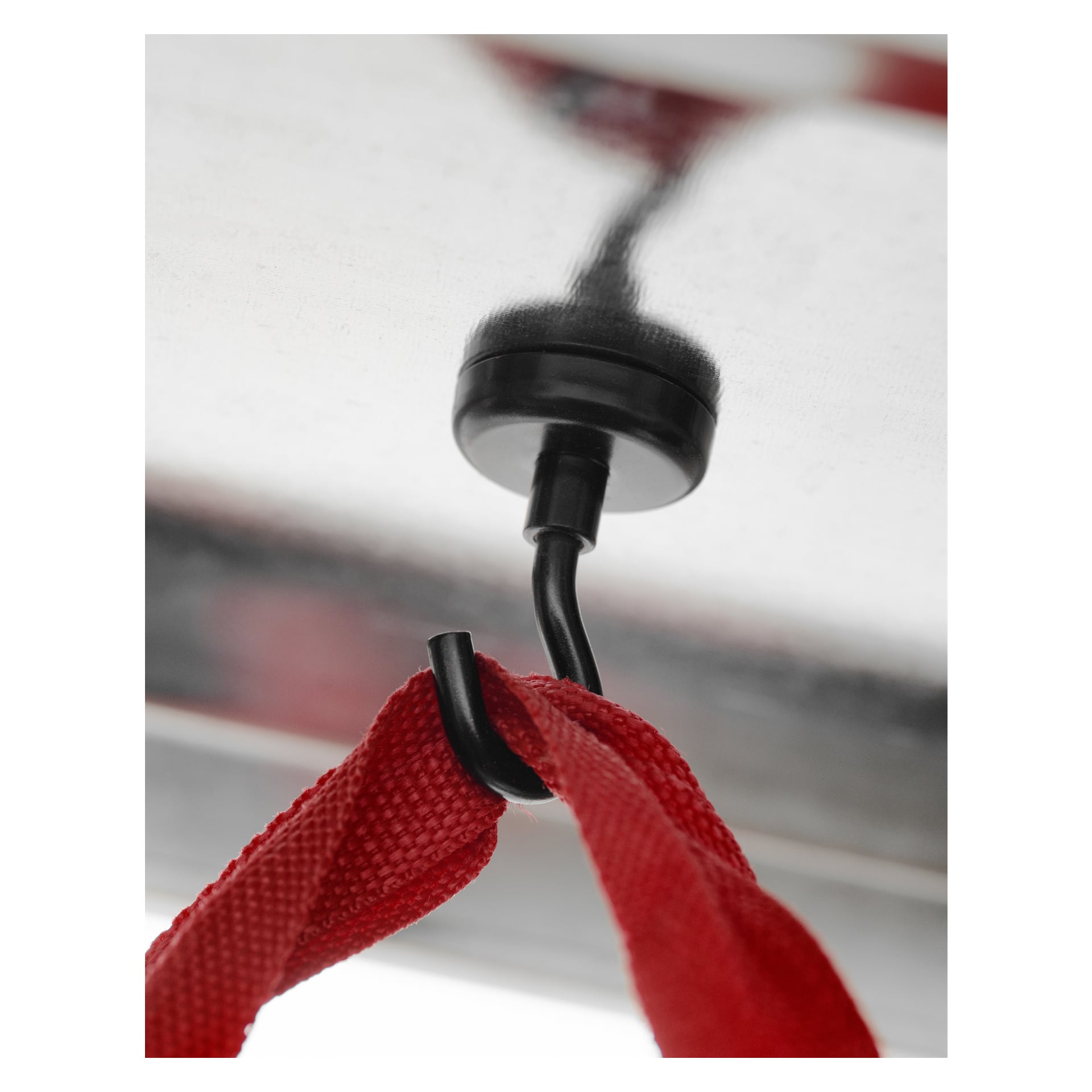 Musta Hard Head -magneettinen koukku "Hooks with magnetic mount 2 pack" -pakkauksesta on kiinnitetty kiiltävään metallipintaan yläpuolella, joka tukee punaisen kangasnauhan lenkkiä. Kuva korostaa sekä koukun että verkon yksityiskohtaisia tekstuureja ja selkeyttä.