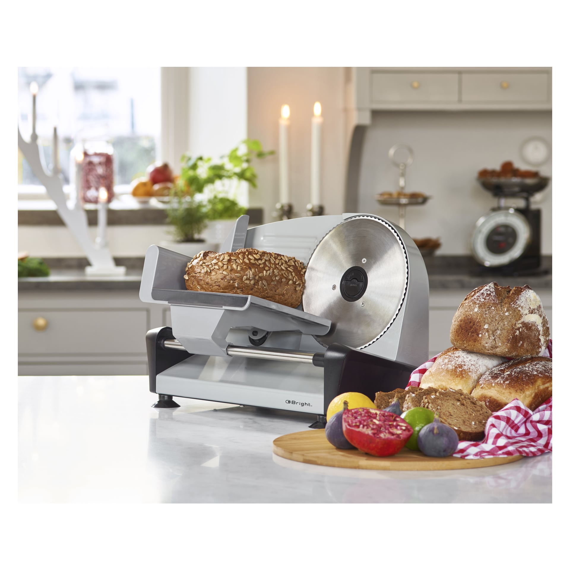 En limpa bröd står ovanpå Bright Slicer i ett solbelyst kök. På bänken i närheten ligger skivat bröd, ett rött och vitt tyg, ett granatäpple, fikon och citroner. Ljus och växter pryder bakgrunden.