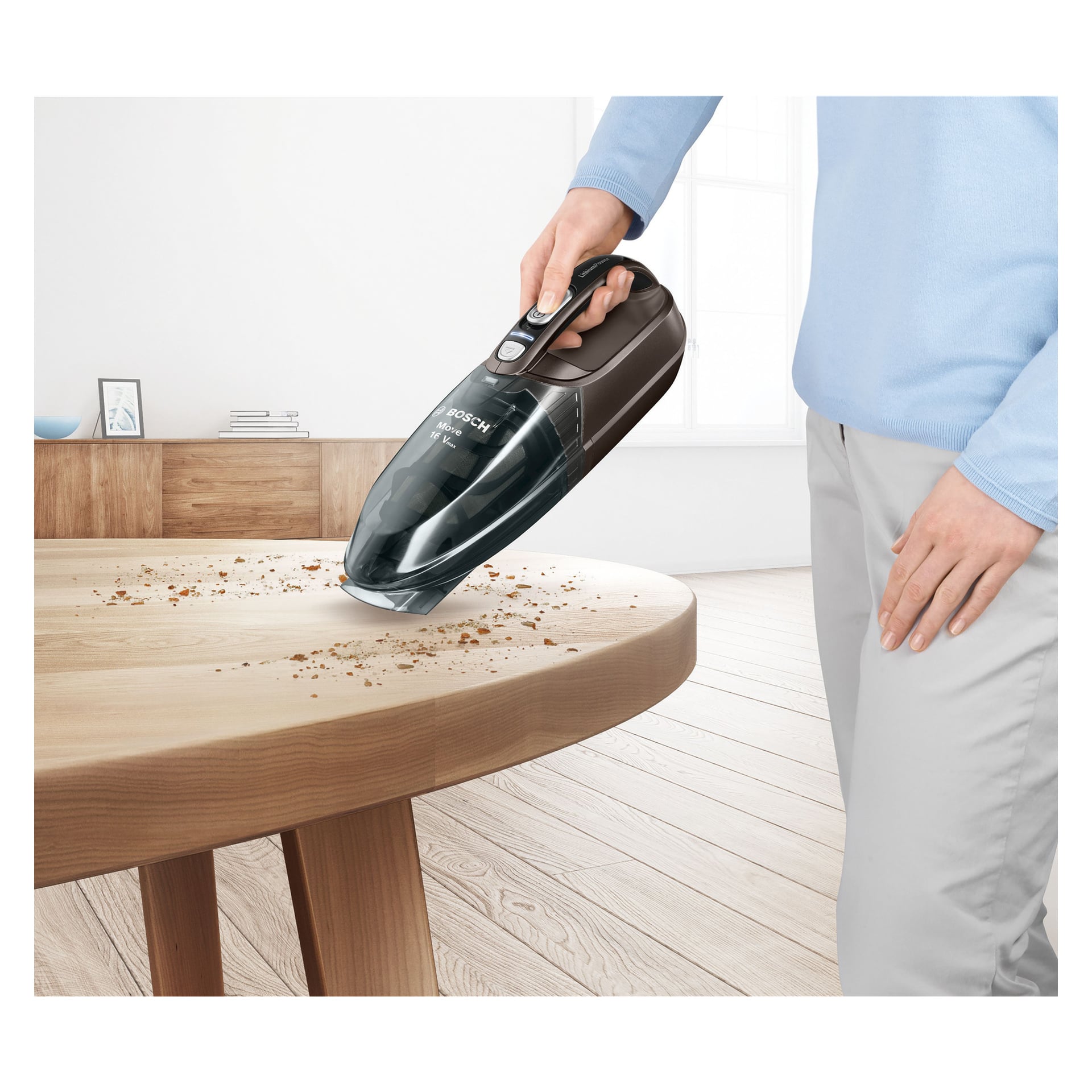 Någon torkar smulor från ett runt träbord i ett ljust, modernt rum med Bosch Home Handheld dammsugare BHN16L.