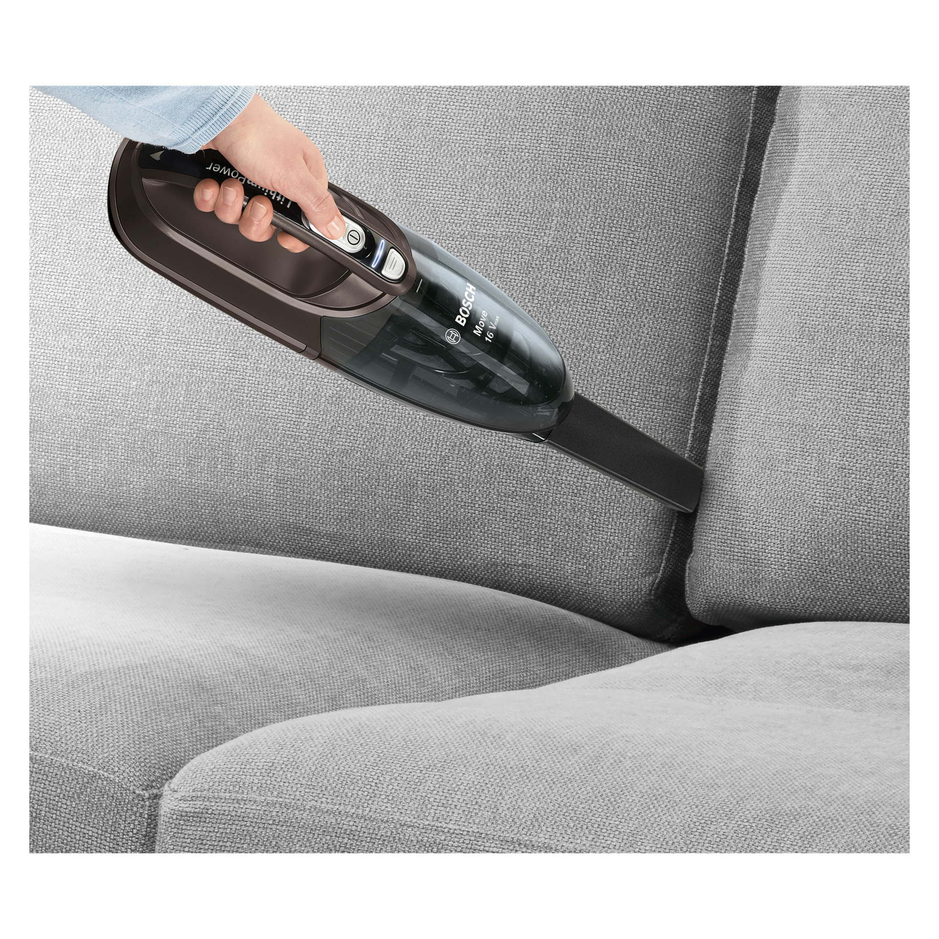 Någon använder Bosch Home BHN16L handhållen dammsugare med ett smalt munstycke för att rengöra mellan två ljusgrå soffkuddar.