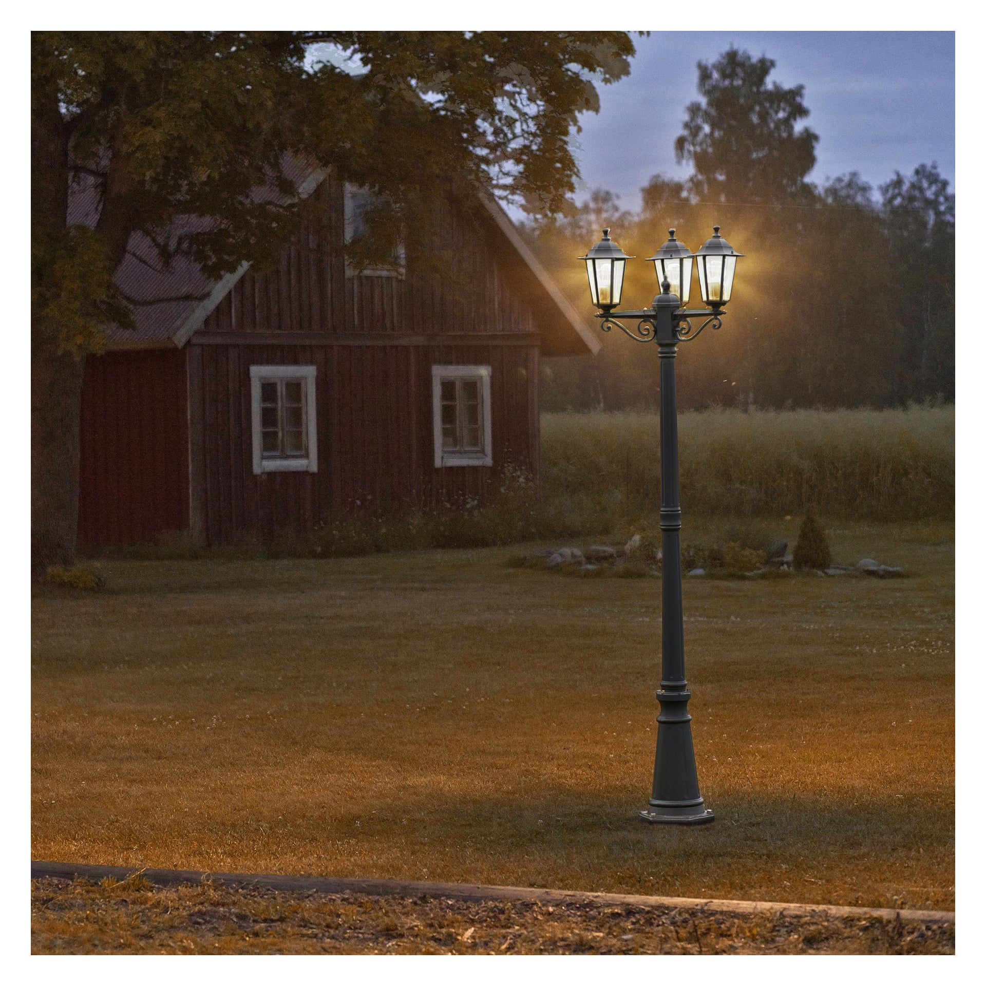 En lyktestolpe fra Anslut kaster lys i skumringen ved siden av et trehus malt rødt med hvite kanter, omgitt av gressplen og trær under en skyet himmel.