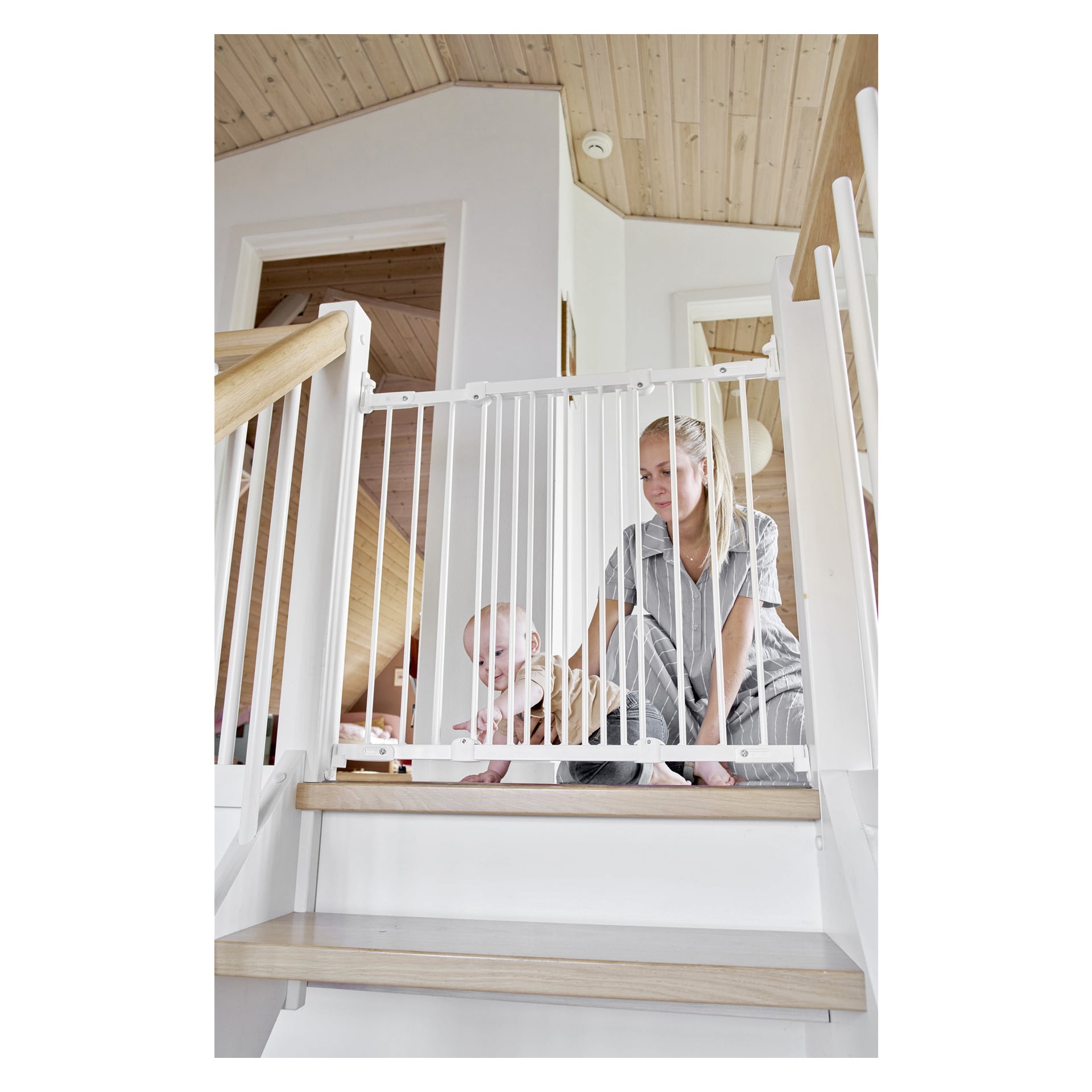 En kvinna och en bebis sitter på golvet bakom Babydan Children's grind Ebba, som är placerad högst upp i en trappa i ett ljust och modernt hem med trätak.