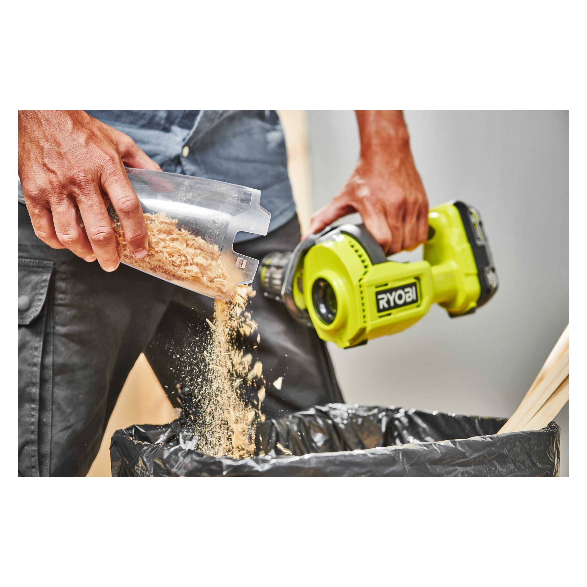 En person häller sågspån från en plastkorg i en soppåse och håller en Ryobi One+ Handhållen dammsugare RHV18-0 i den andra handen.