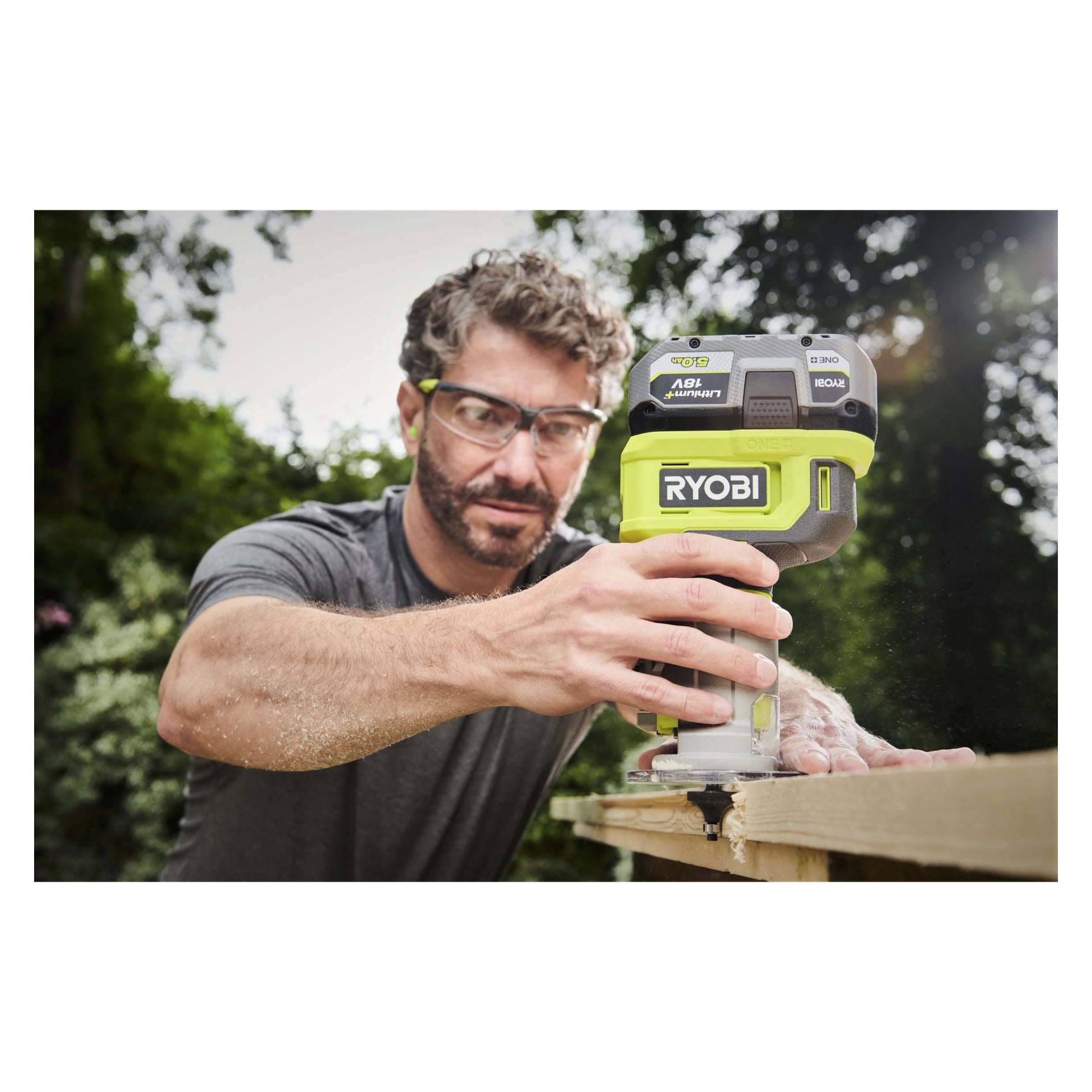En man med skyddsglasögon använder en Ryobi One+ Router RTR18‐0 för att forma en träplanka utanför, med sågspån som sprider sig nära hans hand. Bakgrunden visar suddiga träd och solljus.