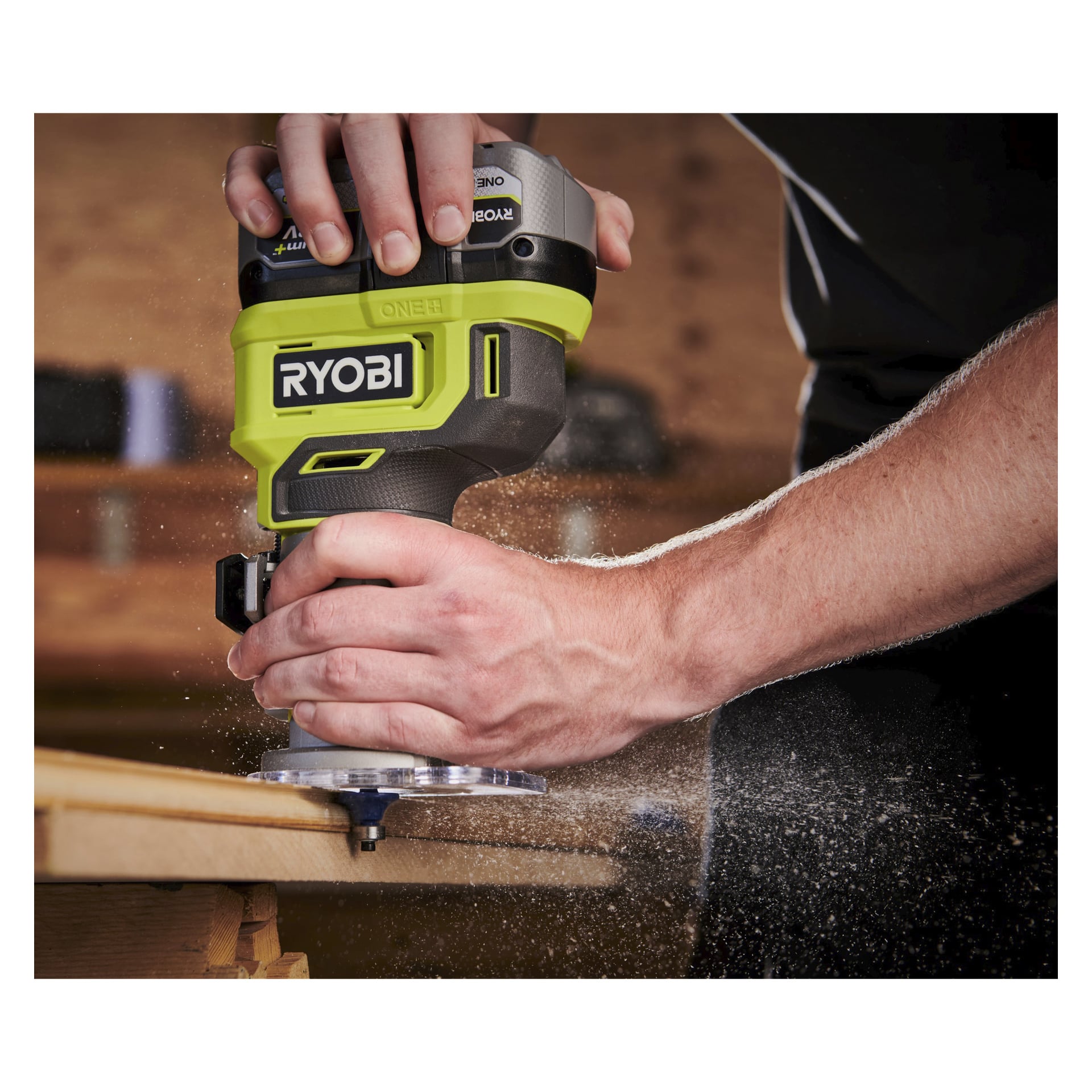 Någon använder en grön och svart Ryobi One+ Router RTR18‐0, formar kanten på en träskiva och producerar sågspån. Bakom dem kan man se en verkstad med suddiga hyllor och olika verktyg.