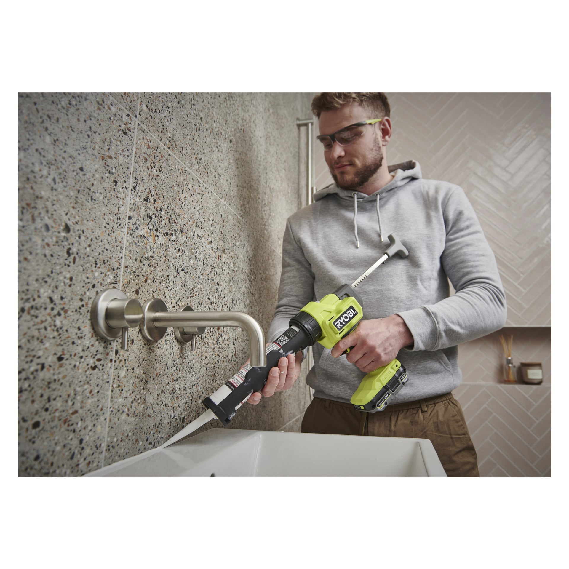 En person i grå hoodie och skyddsglasögon använder en batteridriven Ryobi One+ fogmassa för att applicera tätningsmedel längs kanten på ett badrumshandfat, som har en stenvägg av kakel och en metallkran.
