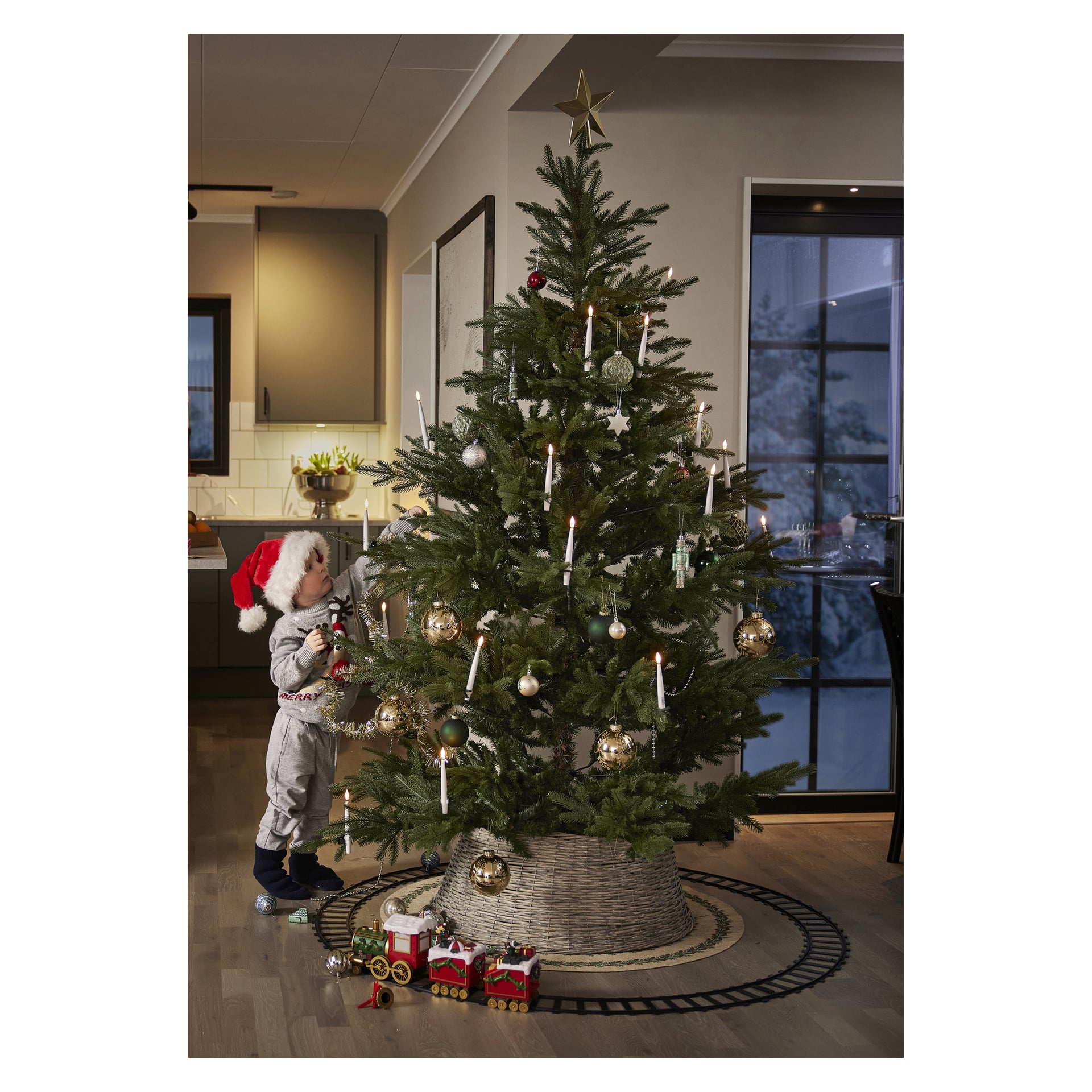 Ett litet barn med tomteluva står bredvid en EKVIP Livslik julgran prydd med ljus och prydnader. Leksakståg går runt trädets bas, med ett kök och ett fönster i bakgrunden.
