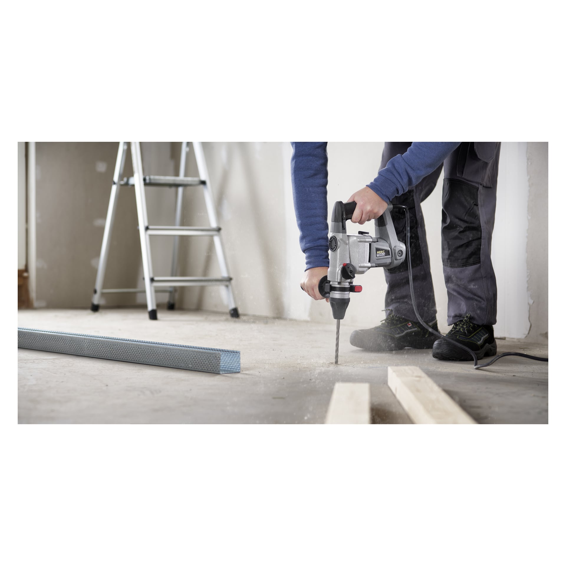 En person klädd i arbetskläder använder en Meec Tools-hammarborr på ett betonggolv och skapar damm när de borrar. I närheten finns träplankor, en metallbalk och en stege.