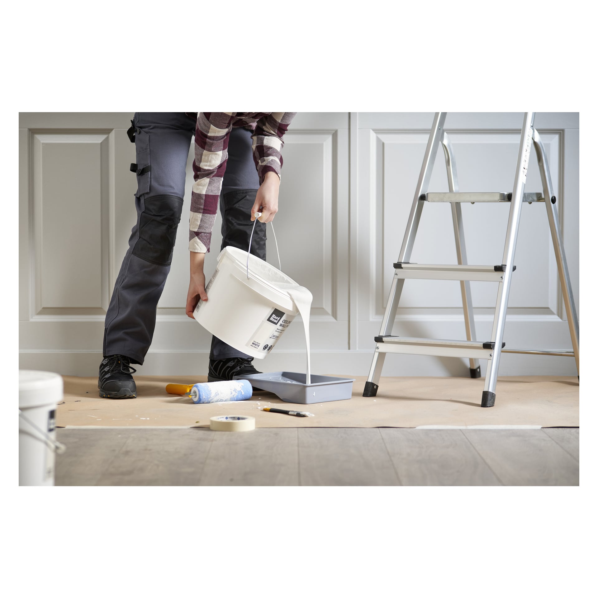 En arbetare häller färg märkt Hard Head Ceiling/Wall-färg i en bricka på ett golv täckt med papper. I närheten finns en roller, tejp, pensel och en metallstege.