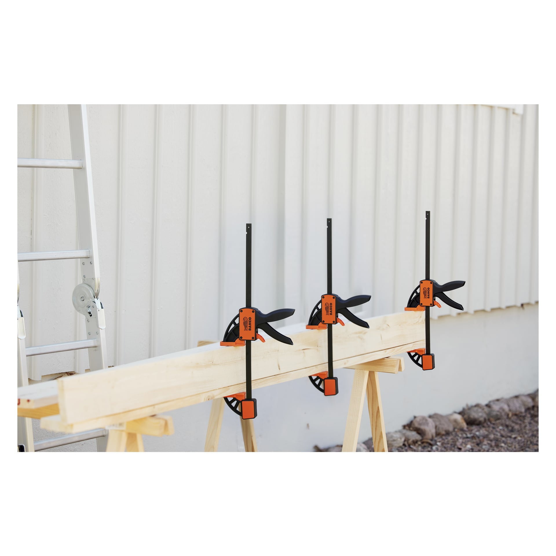 En träplanka fästs vid två såghästar med tre orange och svarta Bahco Quick clamp QC-300A-klämmor. En metallstege dyker upp till vänster, och stenar ligger utspridda på marken i närheten.