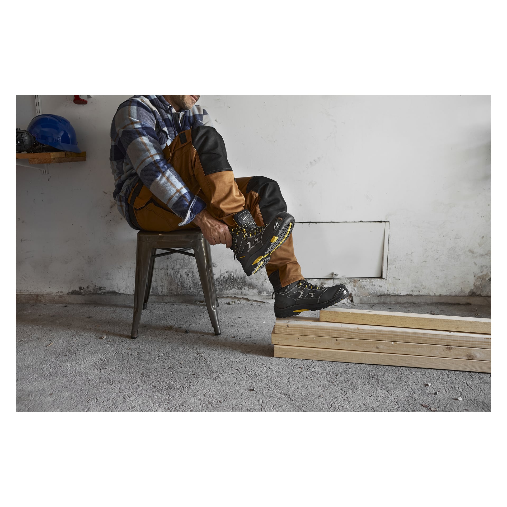 Iklädd arbetskläder sitter personen på en metallpall och justerar sina Bluewear Safety Boots i Vermont. Tre träplankor ligger utspridda på betonggolvet, och en blå säkerhetshjälm vilar på en hylla bakom dem.