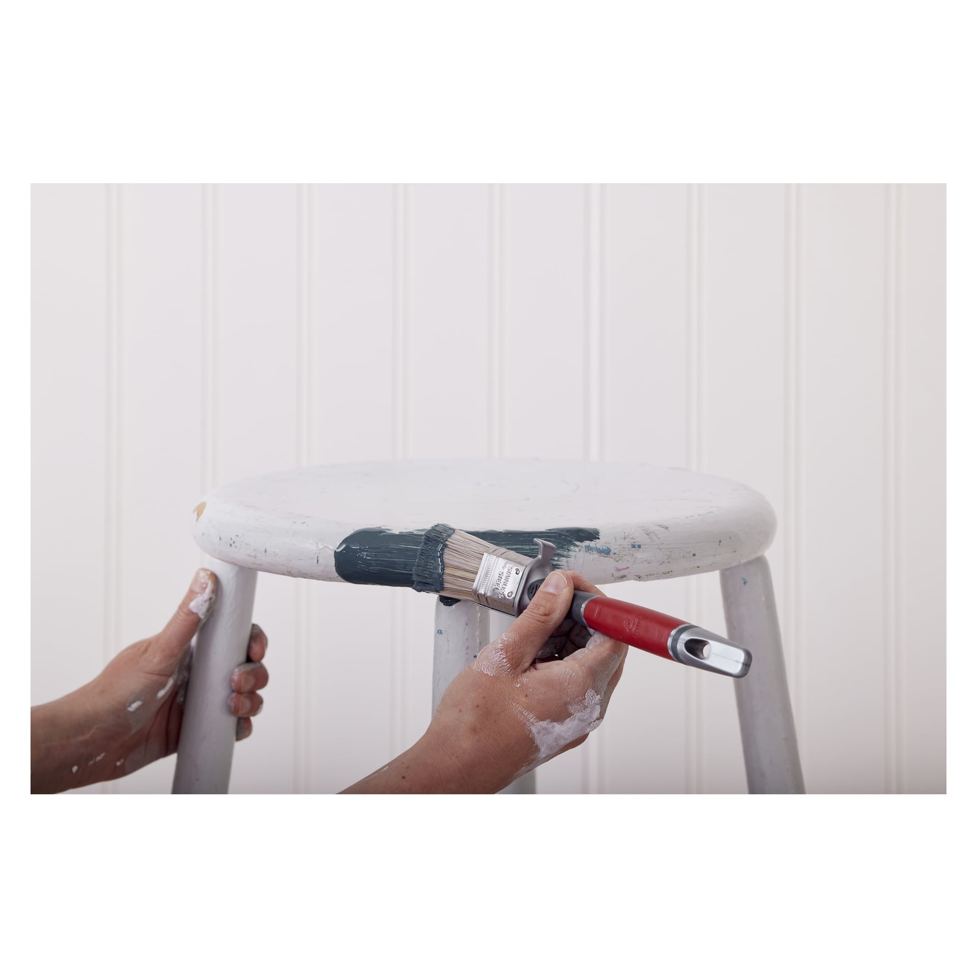 En person använder en Anza Oval Elite-pensel för att applicera mörkgrön färg på kanten av en vit träpall och stabilisera den med ena handen. Vit panel syns i bakgrunden.