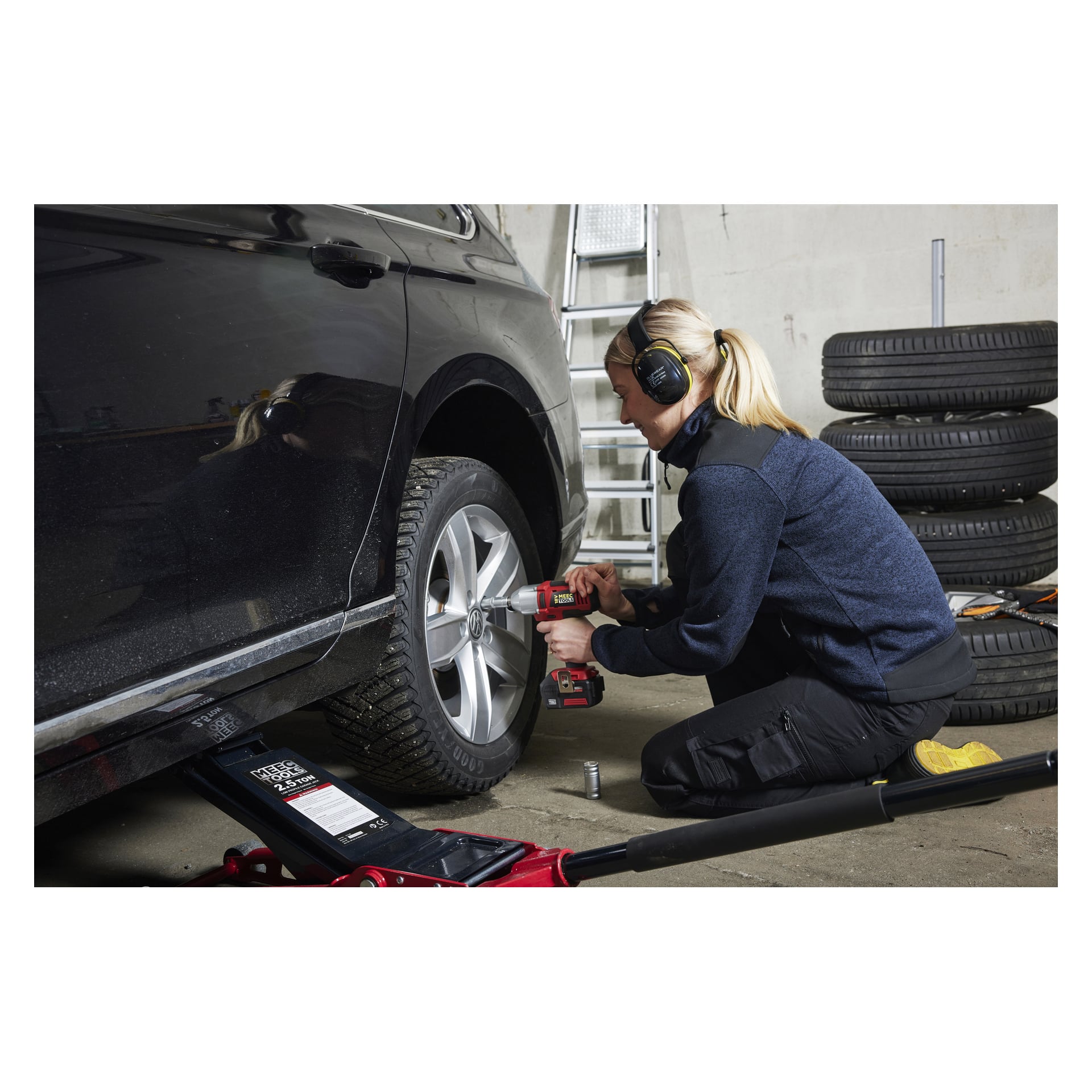 Kuulokkeilla varustettu henkilö käyttää Meec Tools Multiseries IW-18V EASY 100 -akkuiskujakoavainta löysätäkseen auton pyörän mutteria. Autoa tukee tunkki, jossa on pinotut renkaat ja taustalla tikkaat.