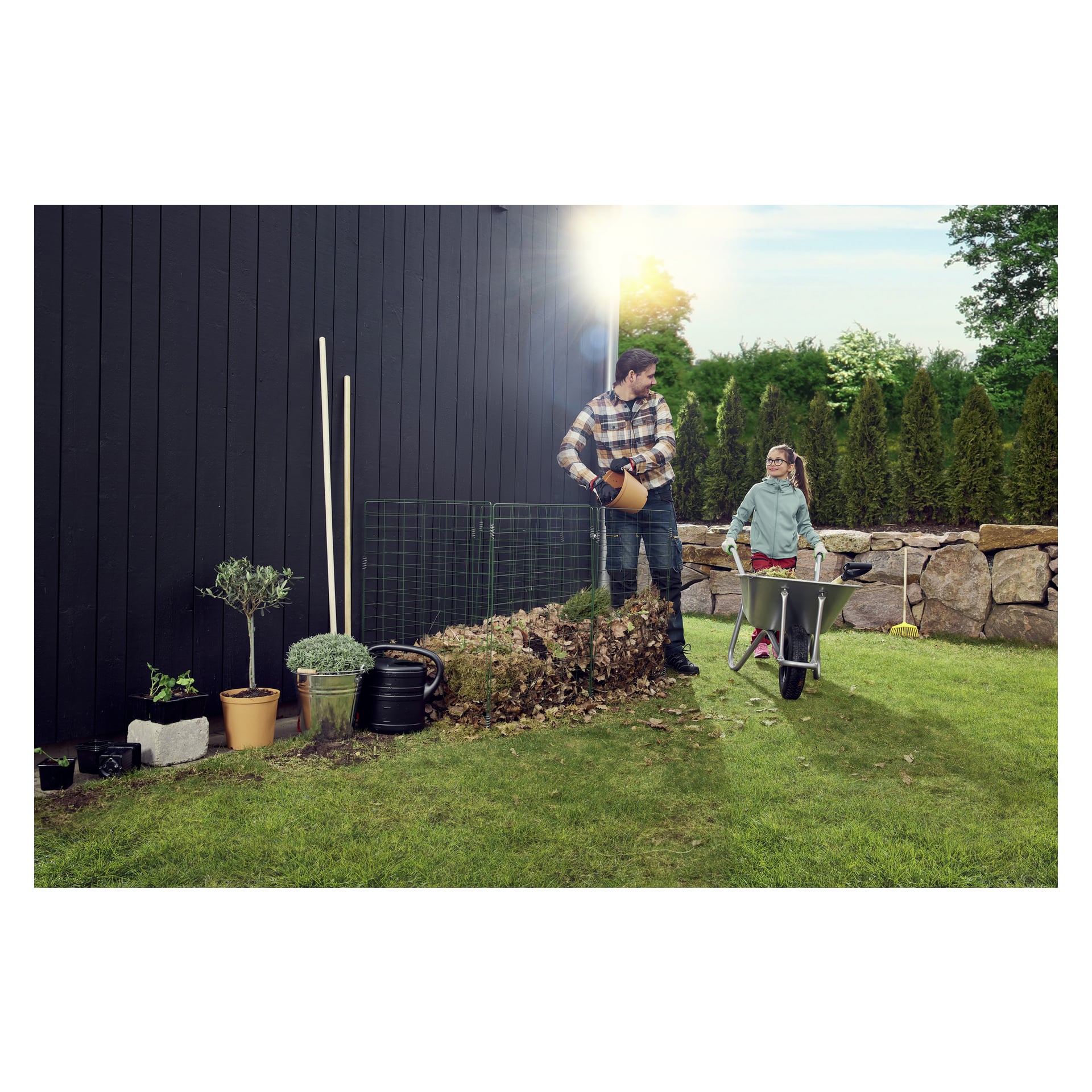En man vattnar växter nära en svart vägg bredvid ett Hard Head Compost-galler, medan en flicka står i närheten med en skottkärra. Olika trädgårdsredskap, krukväxter och en vattenkanna ligger utspridda på gräset. Träd och en stenmur syns i bakgrunden.