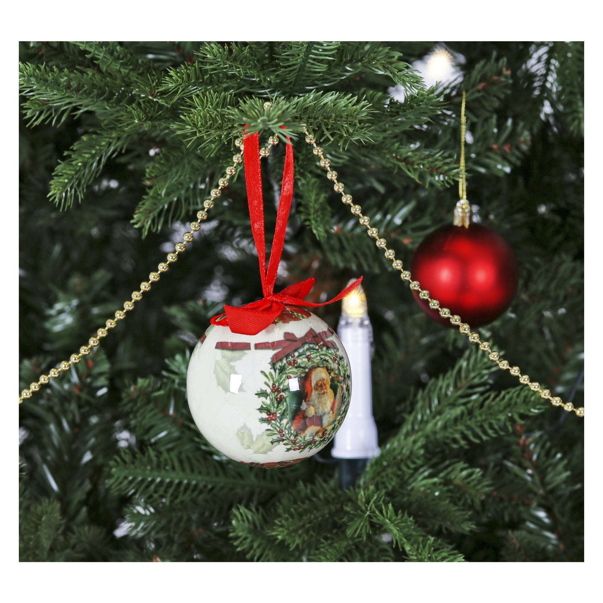 Julgransprydnader från "Christmas tree balls 6-pack" pryder en EKVIP-julgran, såsom en glänsande röd boll och en vit kula med en tomteillustration och rött band, arrangerade bland en guldpärlgirlang. Ett ljusformat vitt ljus lyser mjukt upp de gröna grenarna.