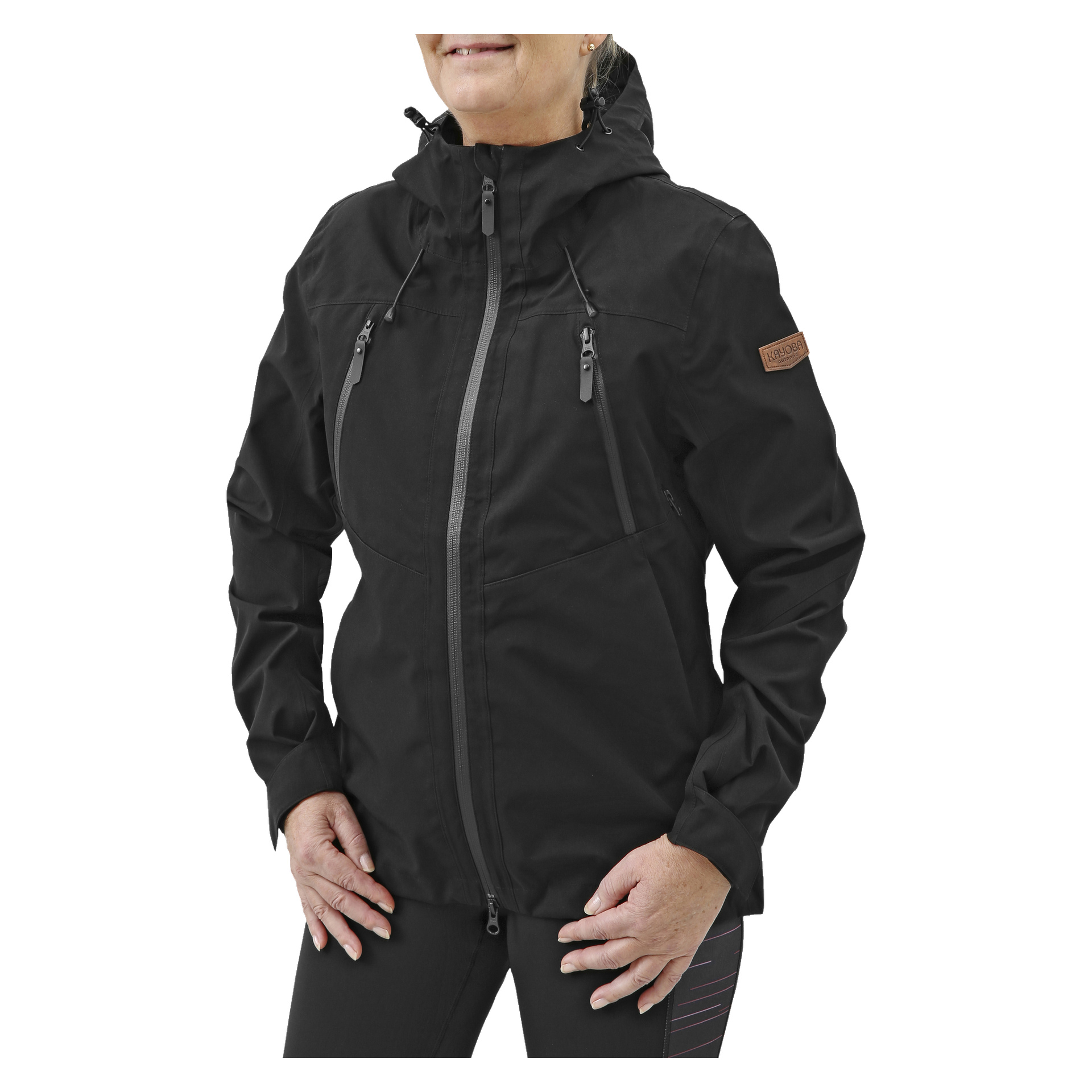 En kvinna står med händerna nära höfterna och visar Kayoba Outdoor Women's shelljacka. Jackan är svart med huva, har flera fickor med dragkedja och har en brun lapp på övre vänstra ärmen.