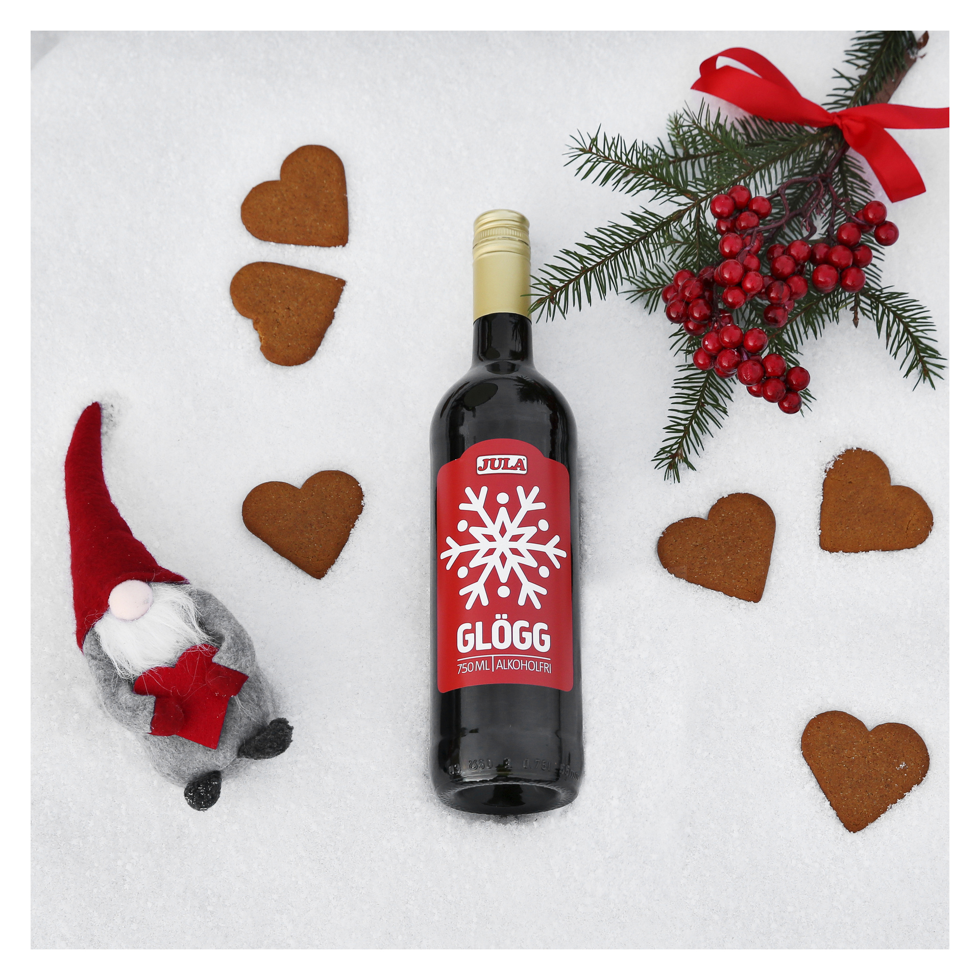 En flaska Sula Glögg står på snön, omgiven av sex hjärtformade Jula-pepparkakskakor, en liten tomtedekoration och en tallgren prydd med röda bär och en röd rosett med ett rött band.