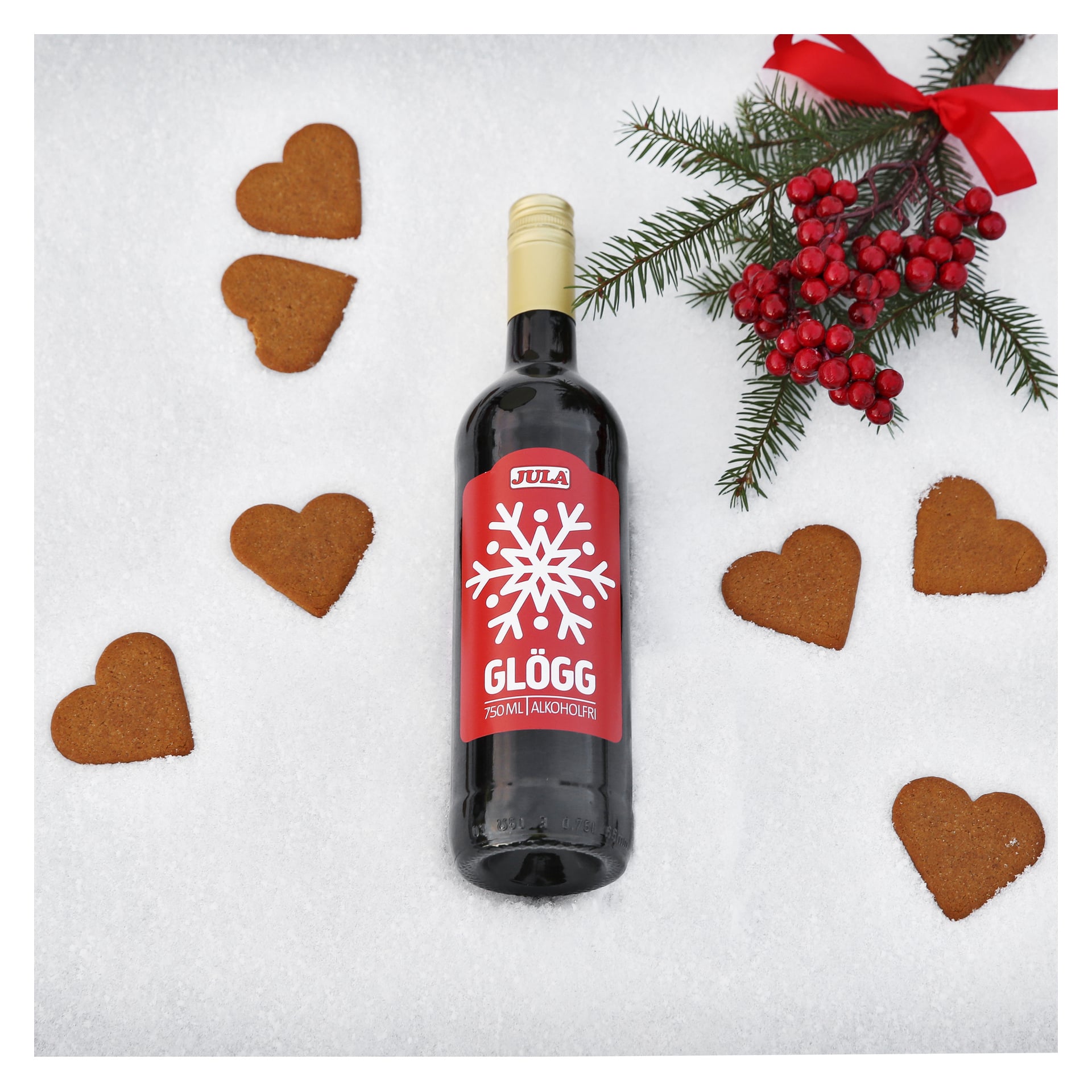En flaska av Julas julglögg står på snötäckt mark, omgiven av sex hjärtformade pepparkakskakor, en tallgren med röda bär och en festlig röd rossett.