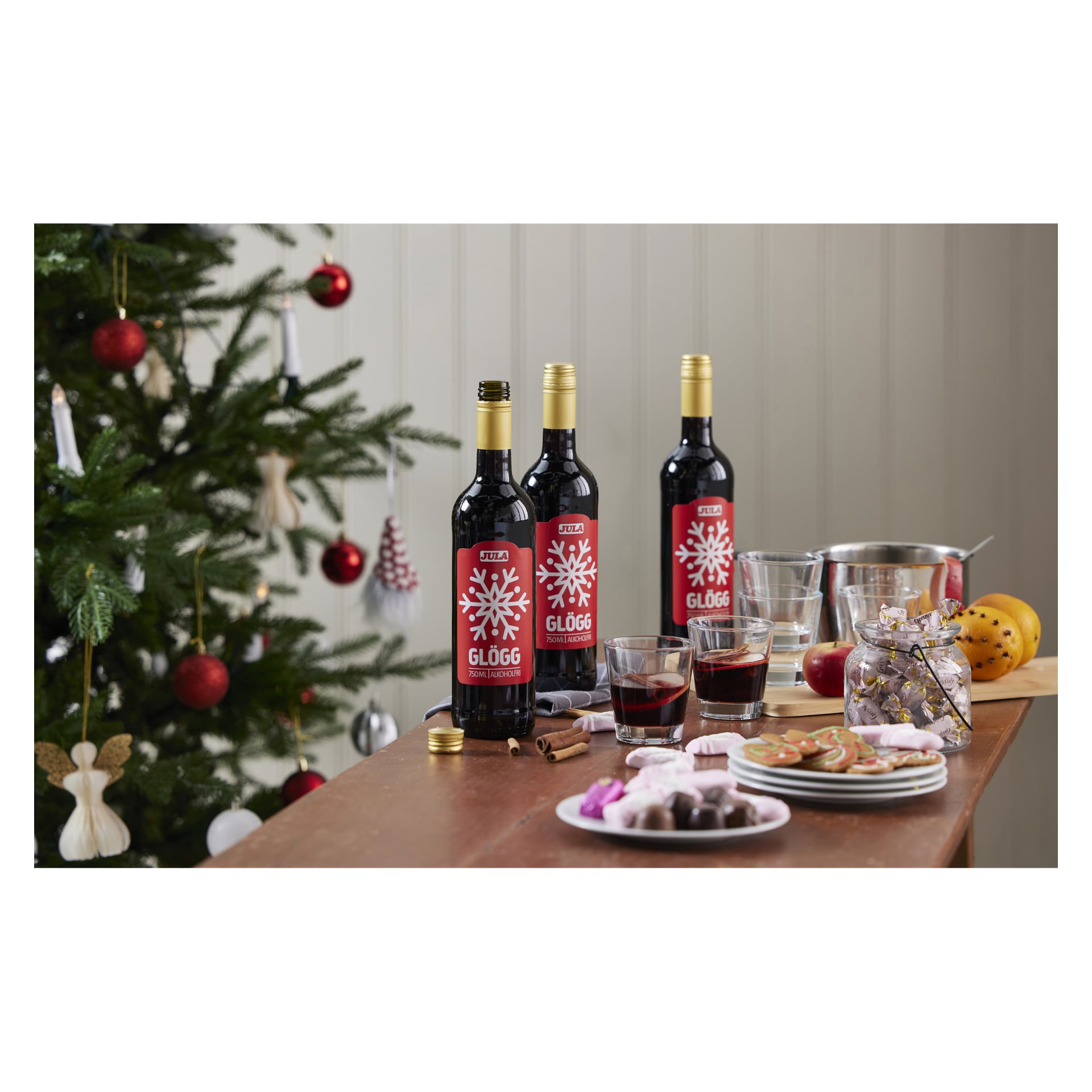 Ett bord är dukat med tre flaskor jul-julglögg, var och en omgiven av glas med kryddad dryck, kakor, olika godis och en mängd festliga dekorationer. I bakgrunden bidrar en julgran dekorerad med röda och vita prydnader till julstämningen.