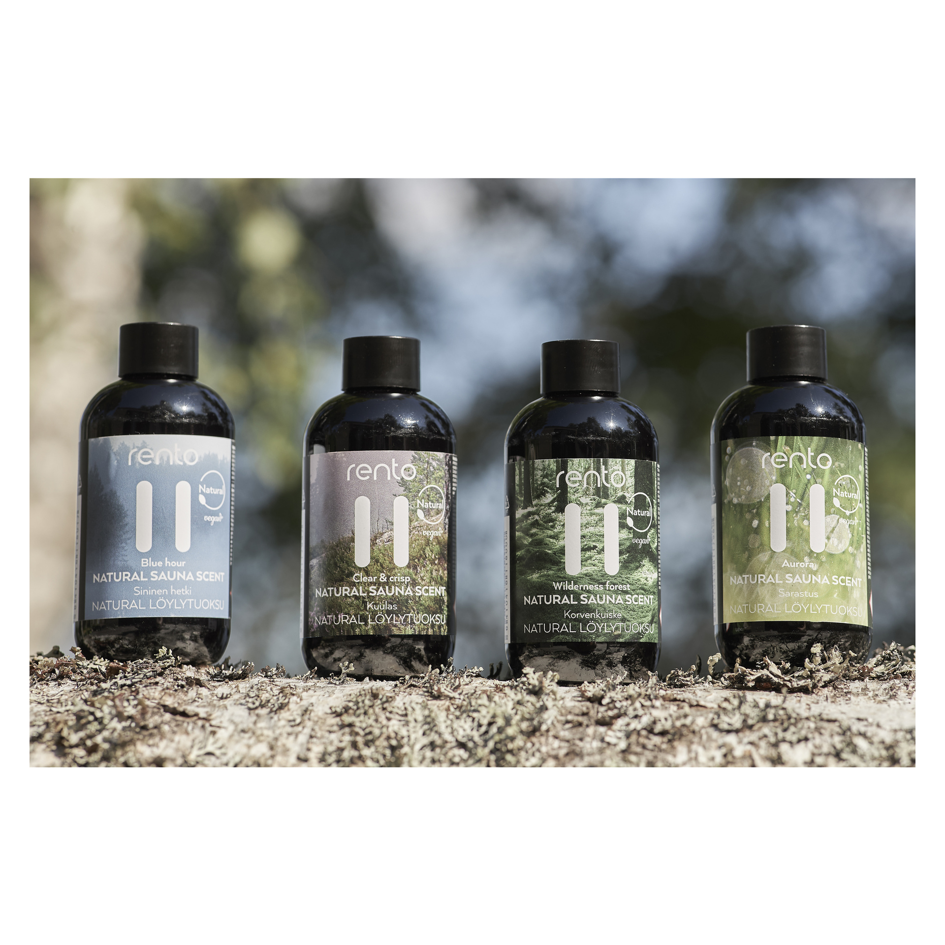 Fyra Rento Sauna aromaflaskor från Natural Sauna Scent-presentpaketet står på en mossbädd utomhus. Varje flaska har en distinkt färgad etikett som anger dofterna: Blå timme, Klar & Krispig, Vildmark och Runt havet.