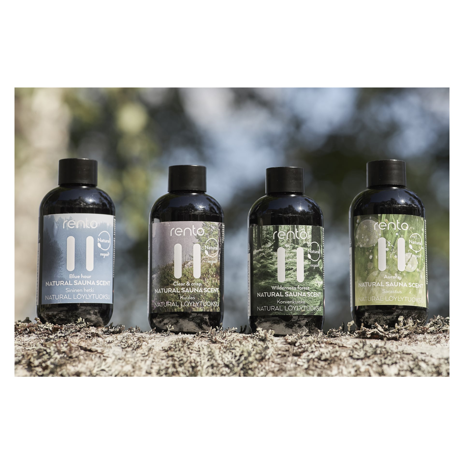 Fyra Rento Sauna aromaflaskor från Natural Sauna Scent-presentpaketet står på en mossbädd utomhus. Varje flaska har en distinkt färgad etikett som anger dofterna: Blå timme, Klar & Krispig, Vildmark och Runt havet.
