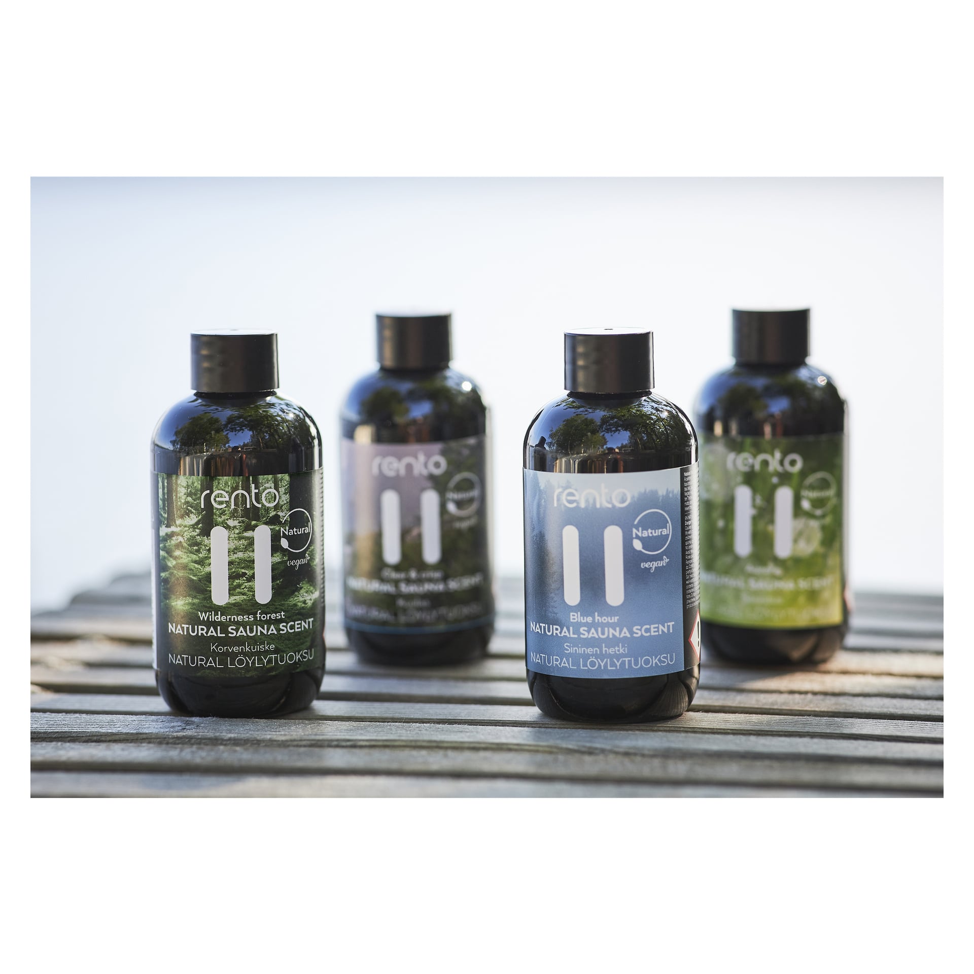 Fyra Rento Sauna Aroma-flaskor från Gift Pack Natural Sauna Scent står utomhus på en träyta. Varje flaska har en distinkt färgad etikett som antyder dess olika doft, med mjukt suddiga träd och himmel i bakgrunden.