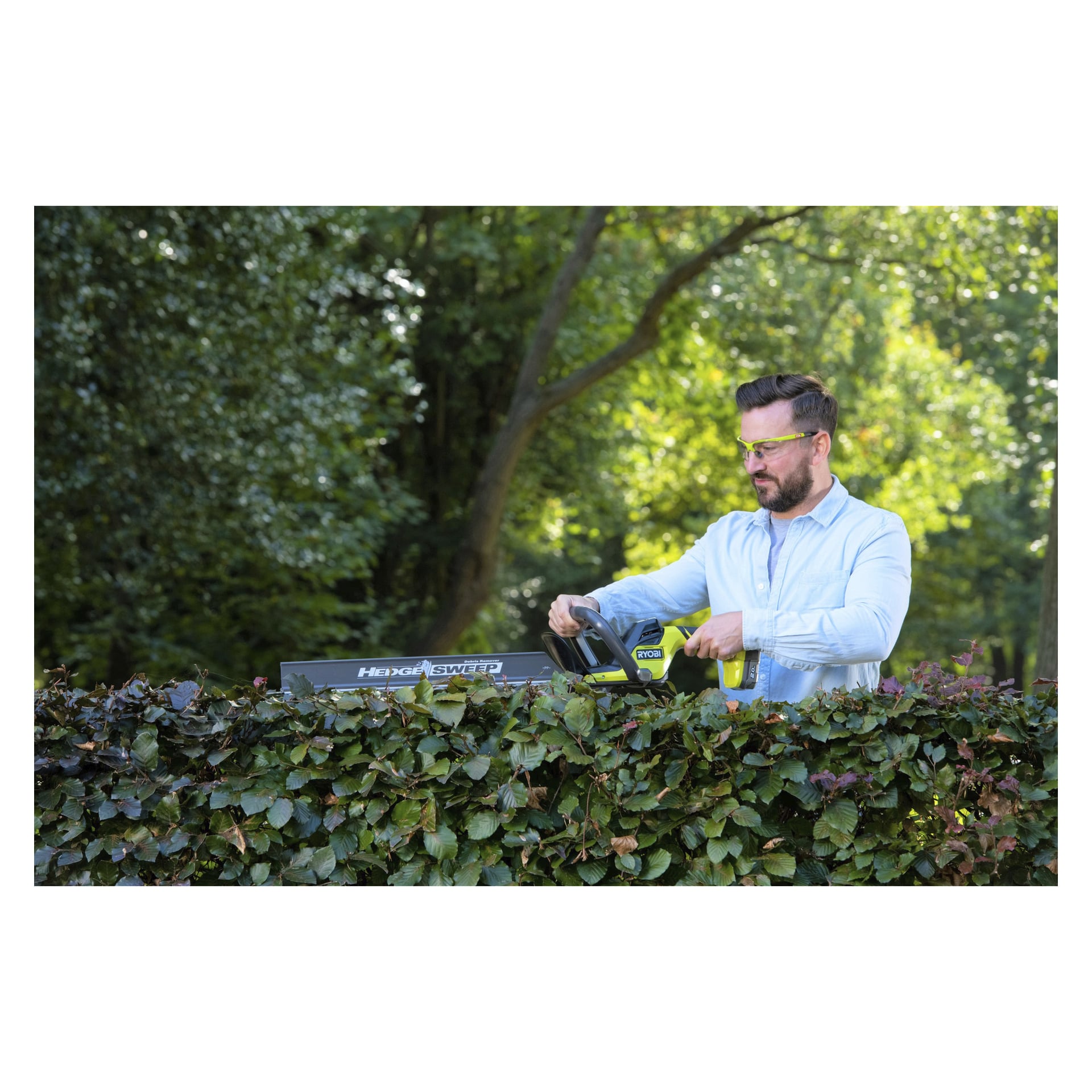 En man med gula skyddsglasögon trimmar en häck med en limegrön Ryobi One+ batteridriven häcksax RY18HT55A-0, som har en häckrensare från Hedge Sweep. Han är ute på en gård, omgiven av frodiga gröna träd.