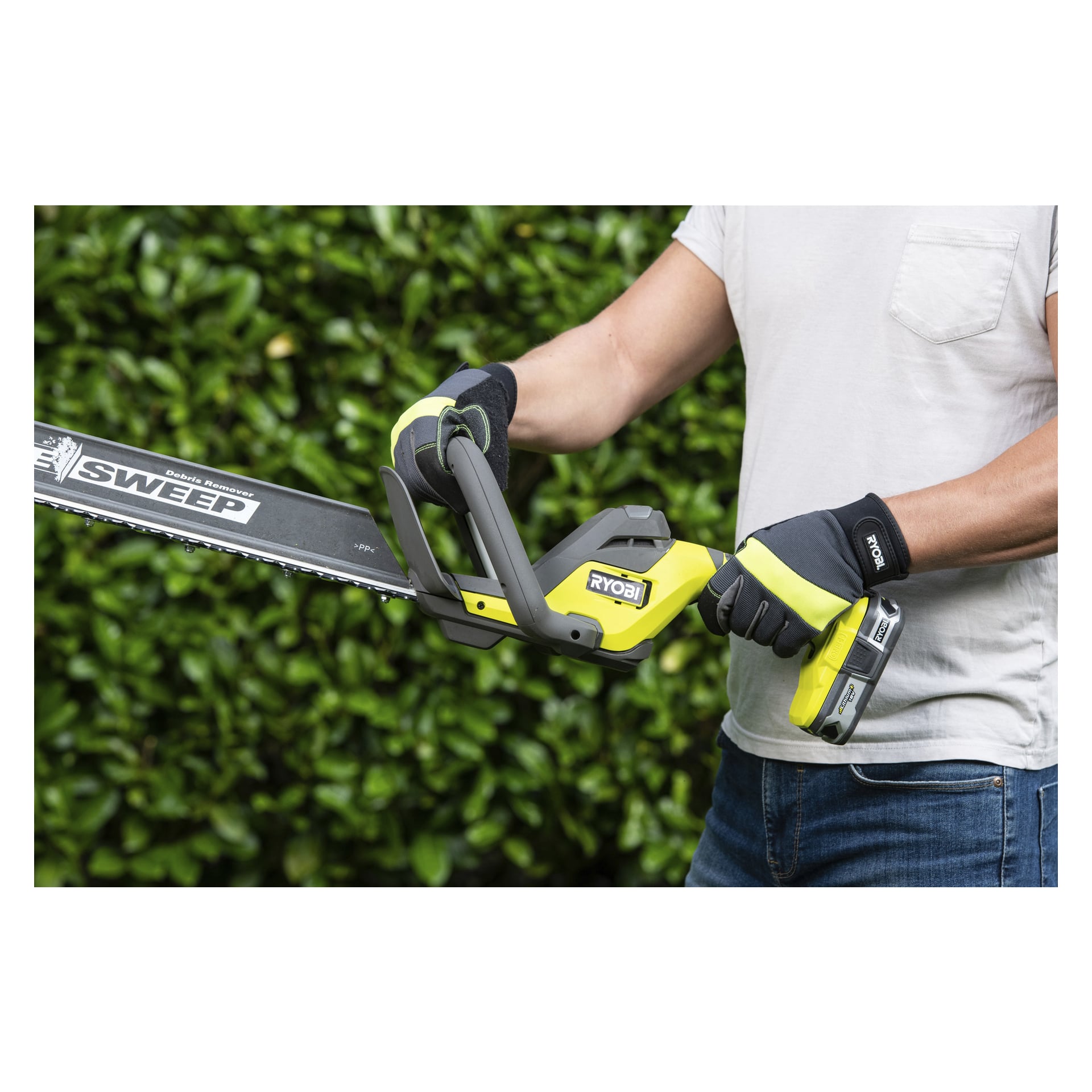 Med handskar och vit t-shirt använder man en Ryobi One+ batteridriven häcksax RY18HT55A-0 utomhus, stående vid frodiga gröna buskar.