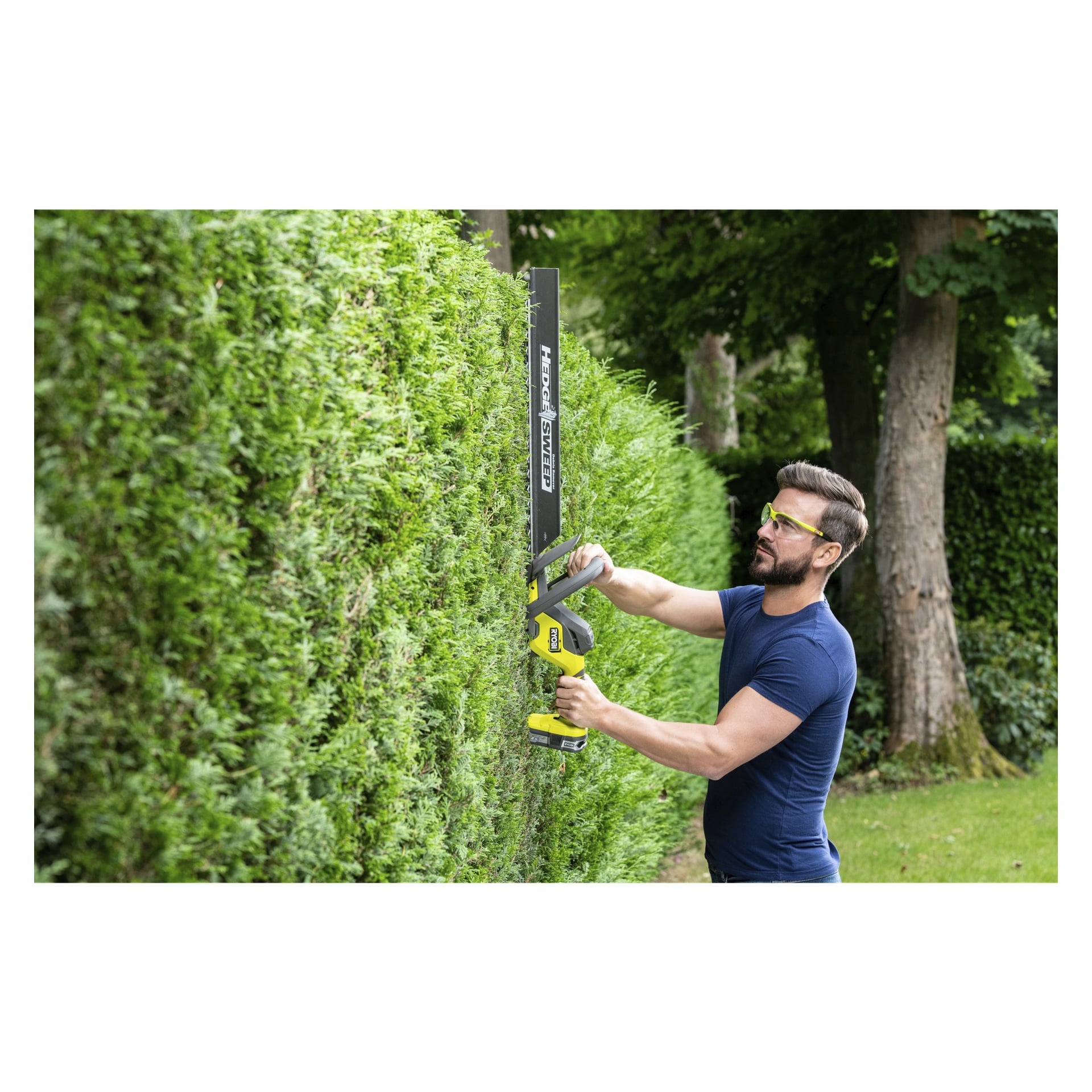 Iklädd en marinblå t-shirt och solglasögon använder en man noggrant Ryobi One+ batteridrivna häcksax RY18HT55A-0 för att forma en hög grön häck i en trädgård omgiven av gräs och träd. Hans koncentration är tydlig när han arbetar.