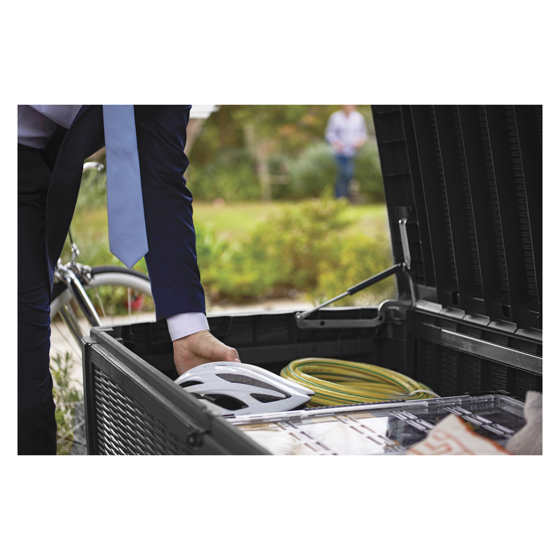 En person som bär kostym och blå slips lägger en vit hjälm i Keter Cushion-lådan Sumatra utomhusförvaringsbehållare, som redan innehåller en gul trädgårdsslang och andra diverse saker. I bakgrunden finns en cykel och en annan otydlig individ.