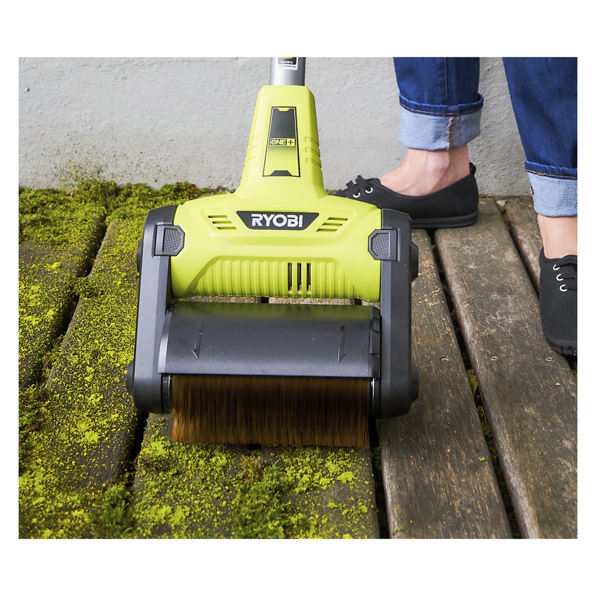 En Ryobi One+ träborste med brunt roterande huvud används för att ta bort grön mossa från trädäck. Fotot visar endast användarens underben och svarta skor.