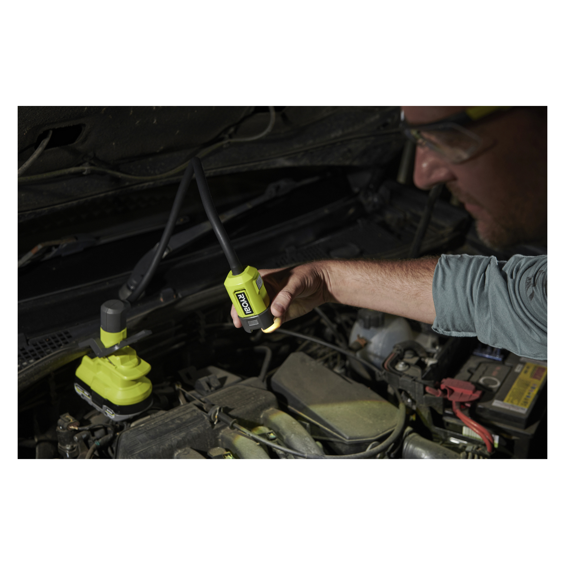En person med vernebriller bruker en Ryobi One+ batteridrevet LED-arbeidslampe med klemme, modell RLCF18‐0, for å lyse motorrommet i en bil med panseret løftet, og fokuserer lysstrålen på deler av motoren.