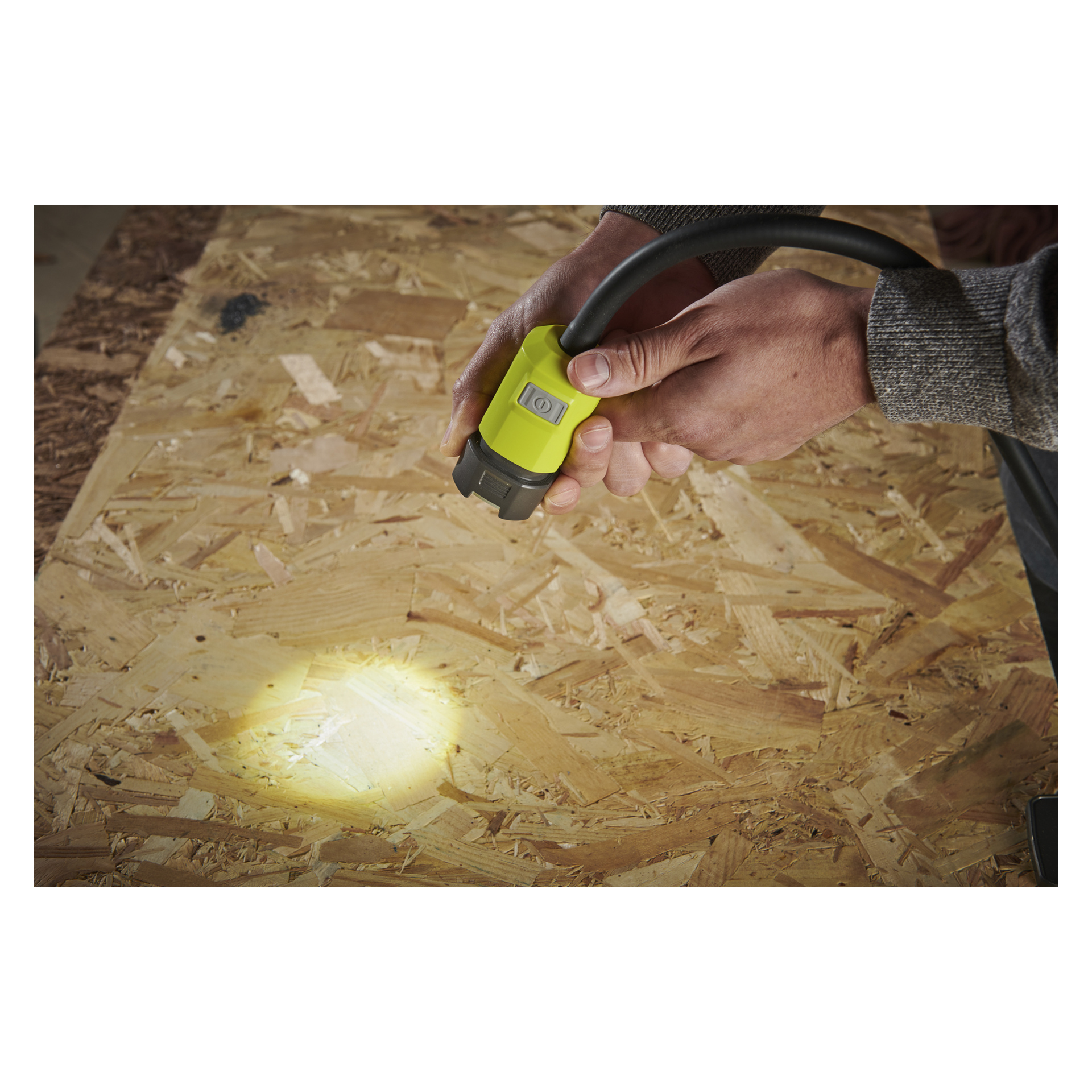 En person med en grå langermet skjorte lyser med Ryobi One+ batteridrevne LED-arbeidslampe med klemme RLCF18-0 på et ubehandlet kryssfinerplate.