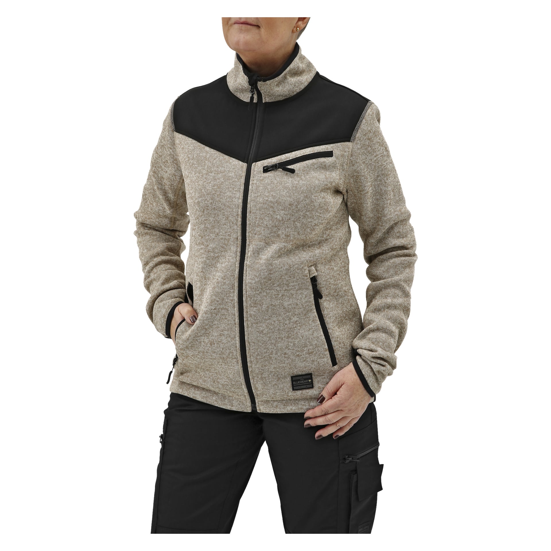 Man bär Women's fleecejacka från Bluewear i beige med svarta detaljer, tillsammans med svarta byxor. Händerna stoppas ner i jackfickorna.