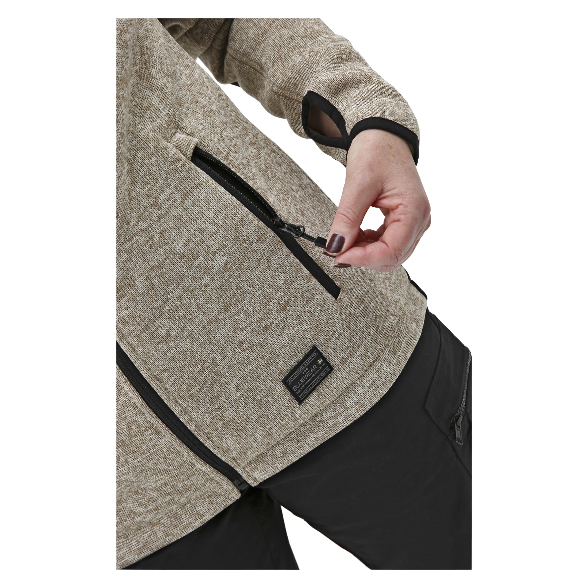 Eine Person in schwarzer Hose und einer beigen Bluewear Fleecejacke öffnet eine Seitentasche. Die Jacke hat einen schwarzen Reißverschluss, einen Daumenloch-Ärmel und ein kleines rechteckiges Etikett neben der Tasche.