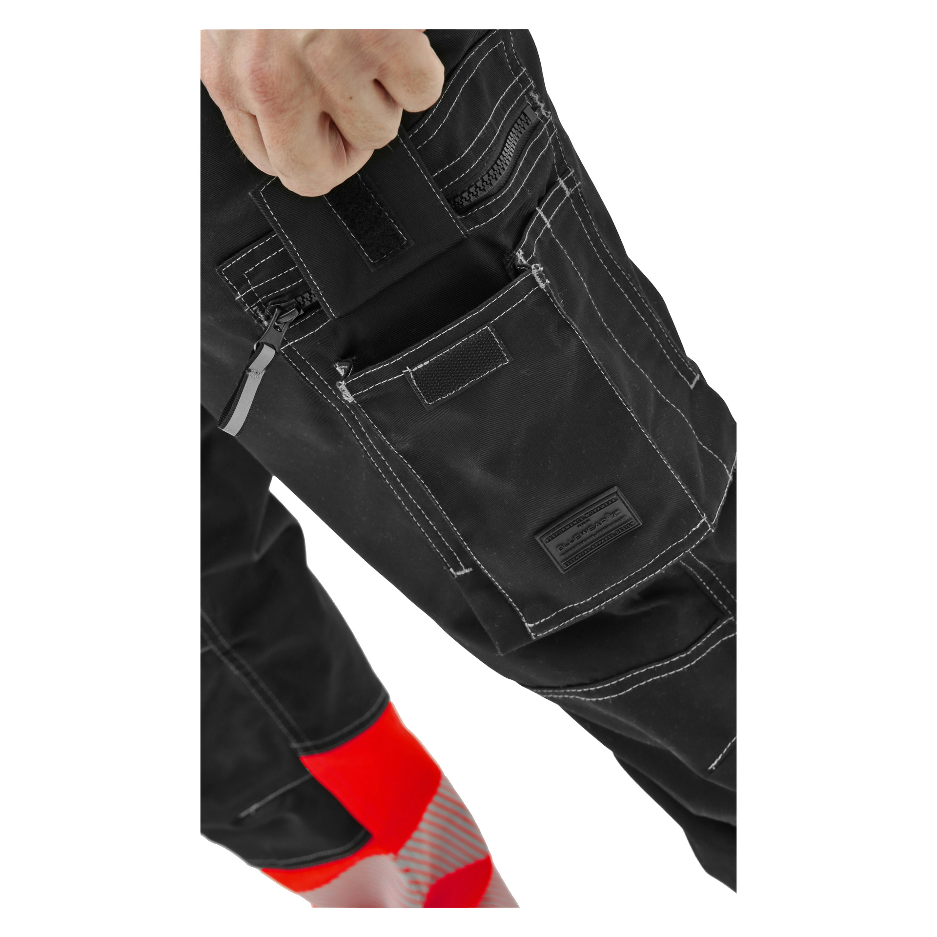 Nahaufnahme einer Person, die eine schwarze Arbeitshose mit weißen Nähten trägt. Zu sehen sind Taschen mit Reißverschluss und Klettverschluss sowie eine Hand und ein Teil eines Beins mit einem rot-weiß gestreiften Detail an der Unterseite.