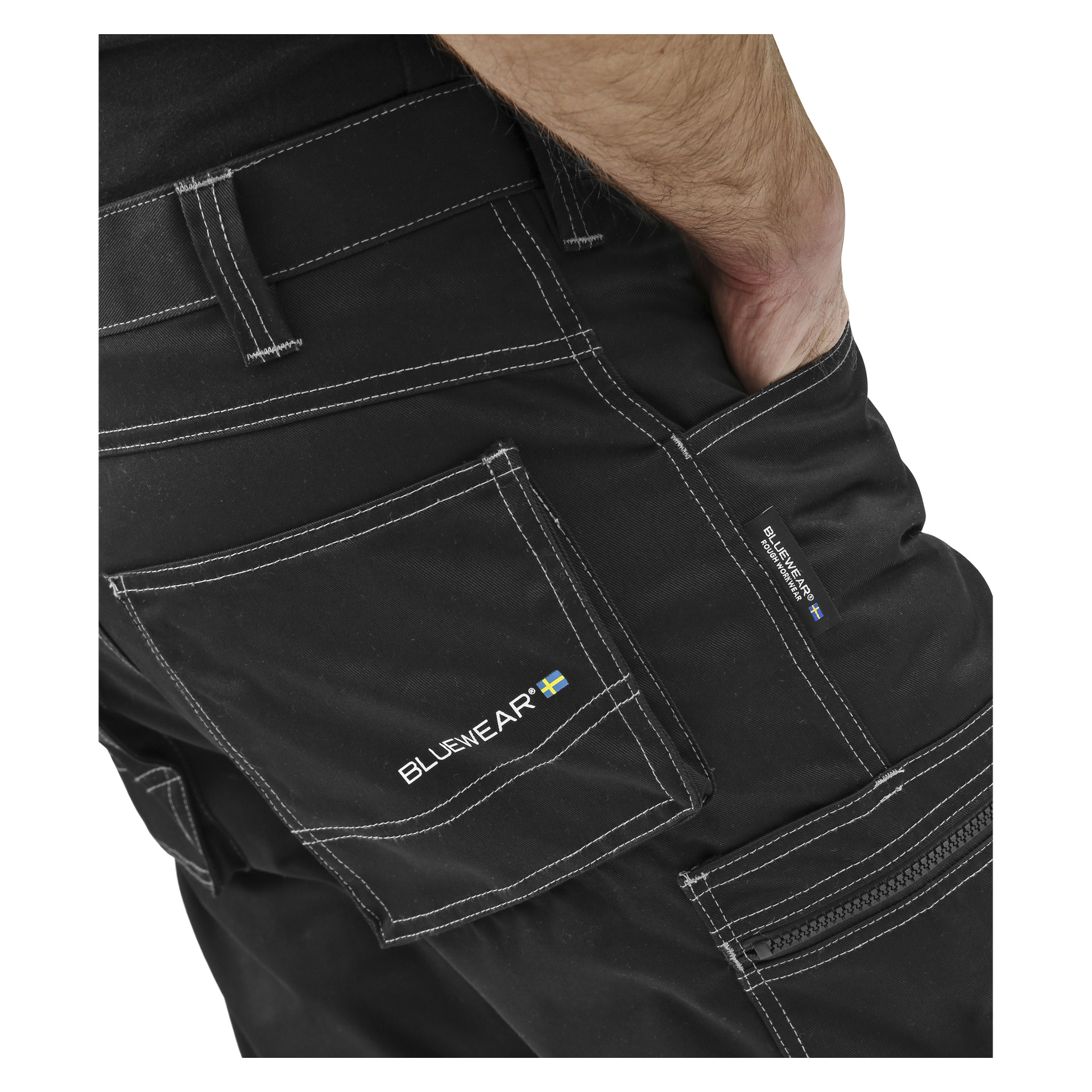 Eine Person trägt schwarze Hi-Vis-Bluewear-Hosen ohne Nageltaschen, mit weißen Nähten und steht mit der rechten Hand in der Tasche. Bluewear und eine schwedische Flagge erscheinen auf der Rücktasche und dem Seitenetikett.
