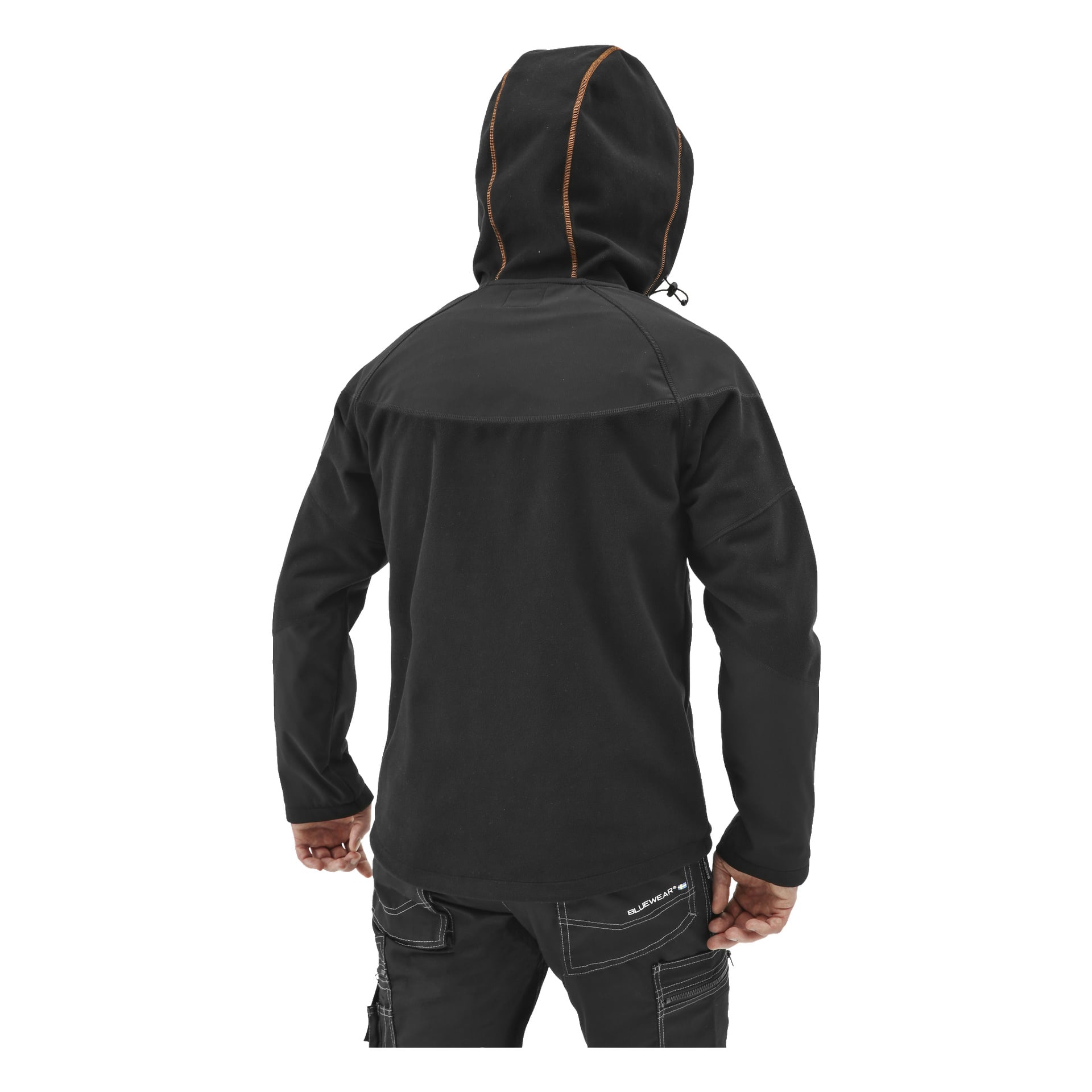 En person står med ryggen mot betraktaren, klädd i en svart Bluewear Pro Workwear herrjacka med vindtät fleecejacka och bruna sömdetaljer, tillsammans med svarta byxor. Deras händer vilar naturligt vid sidorna och är delvis synliga.