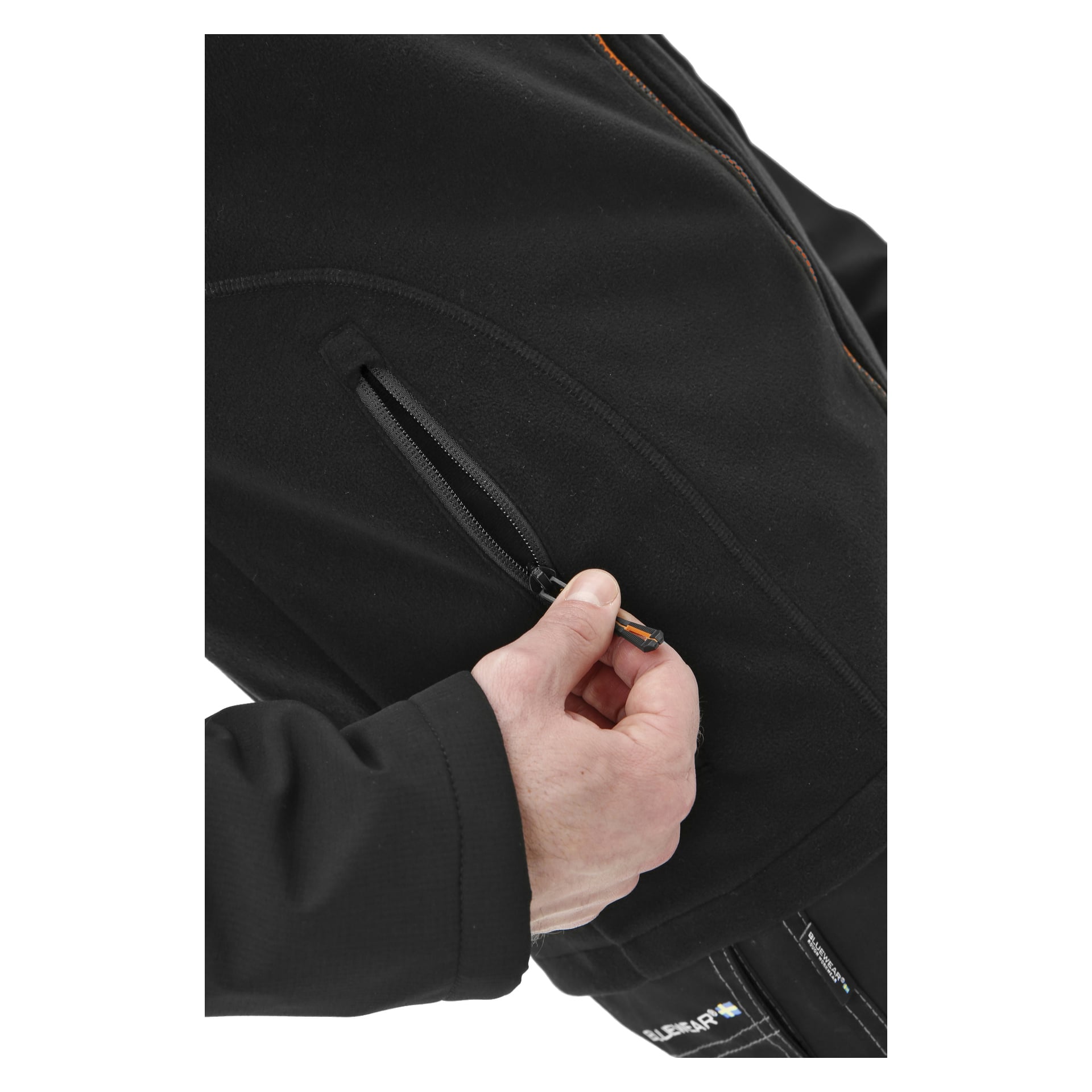 En person använder sin högra hand för att öppna fickan på en Bluewear Pro Workwear herrjacka i vindtät fleecejacka, med betoning på handen, dragkedjan och fickan samt en del av jackans framsida och ärm synlig.